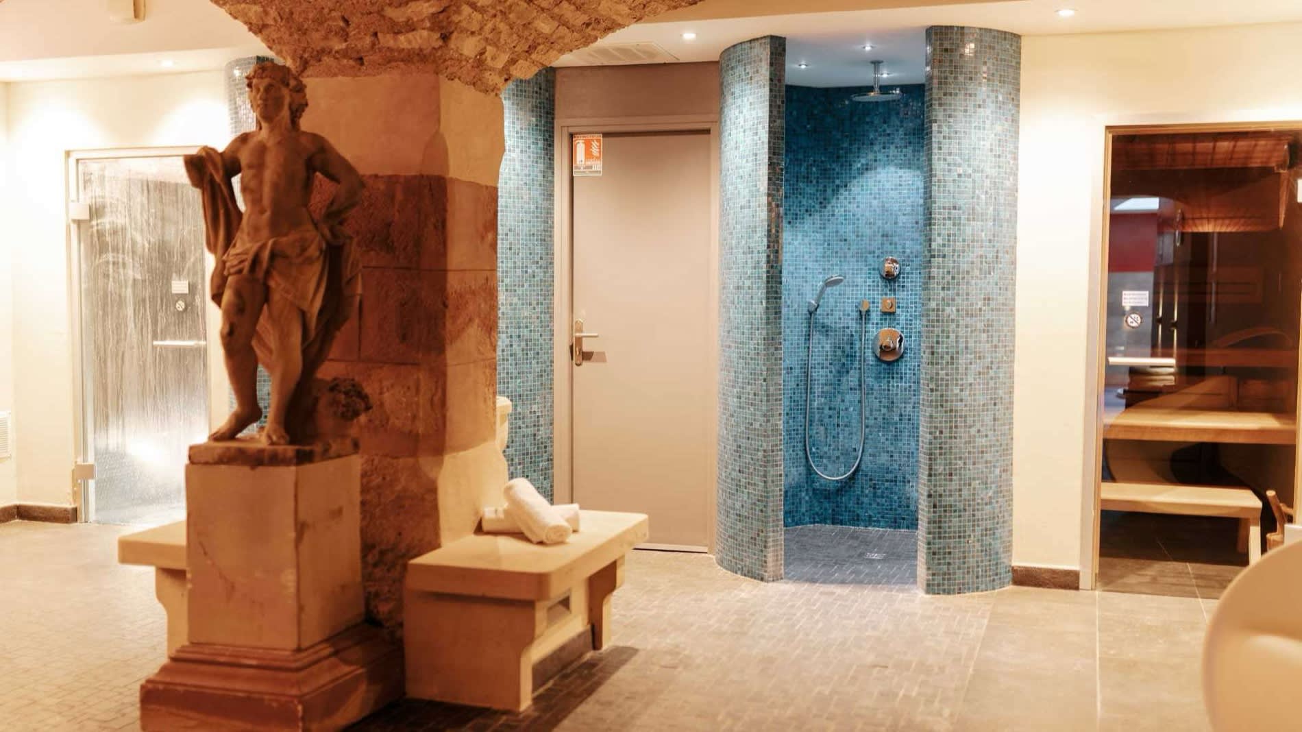 Le spa voûté de l'hôtel Le Bouclier d'Or à Strasbourg, avec sa statue en grès des Vosges, sa douche en mosaïque bleue et l'entrée du sauna