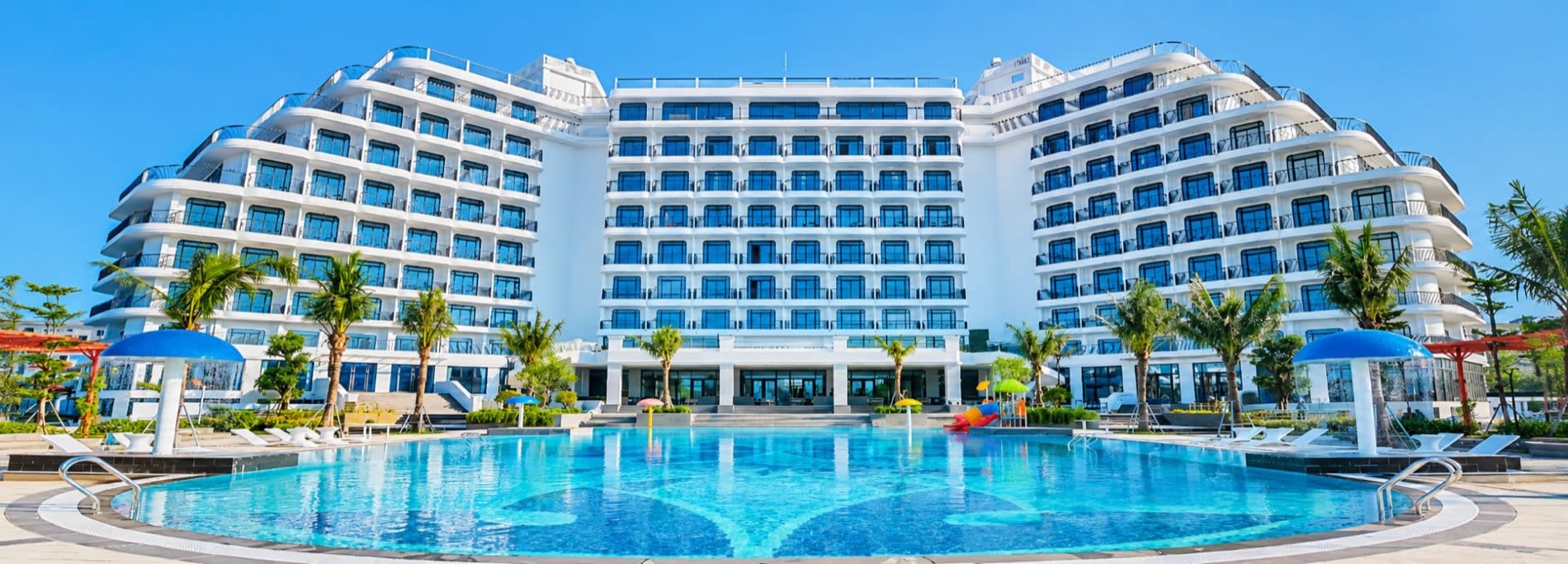 Facade blanche du Tra Co Long Beach Hotel & Resort a Mong Cai vue depuis la grande piscine exterieure bordee de palmiers