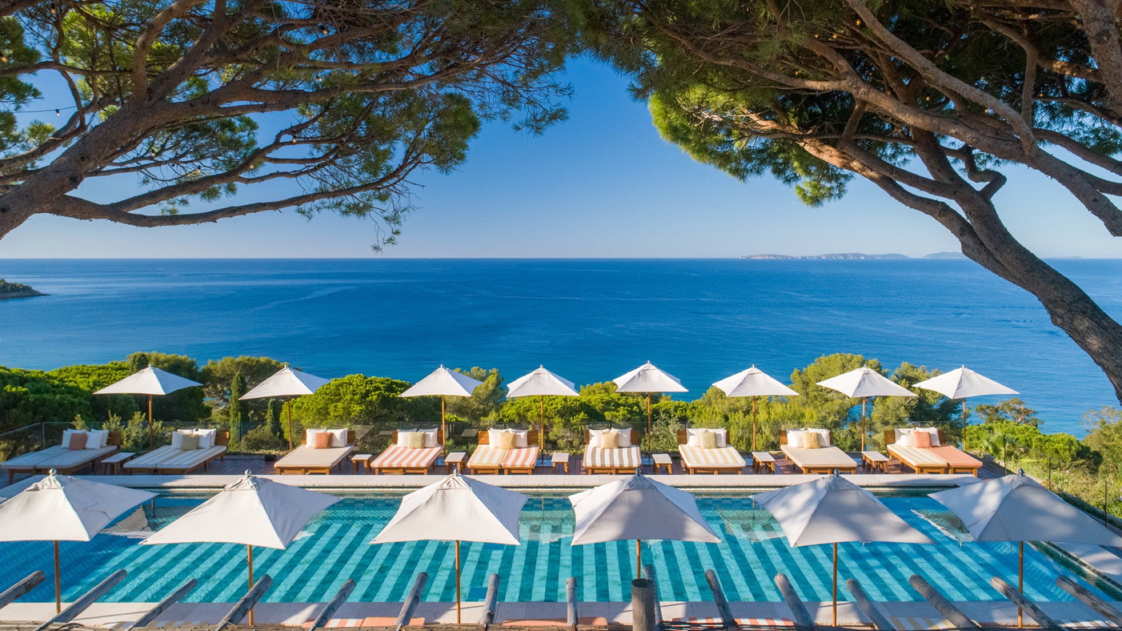 Infinity pool at a luxury resort with ivy-covered stone arches, sun loungers, and a clock tower against a blue sky reflection in the water.