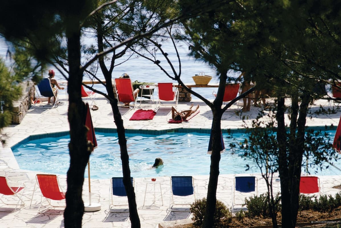 Woman in a bikini walking on a pool deck overlooking the sea, carrying a hat.