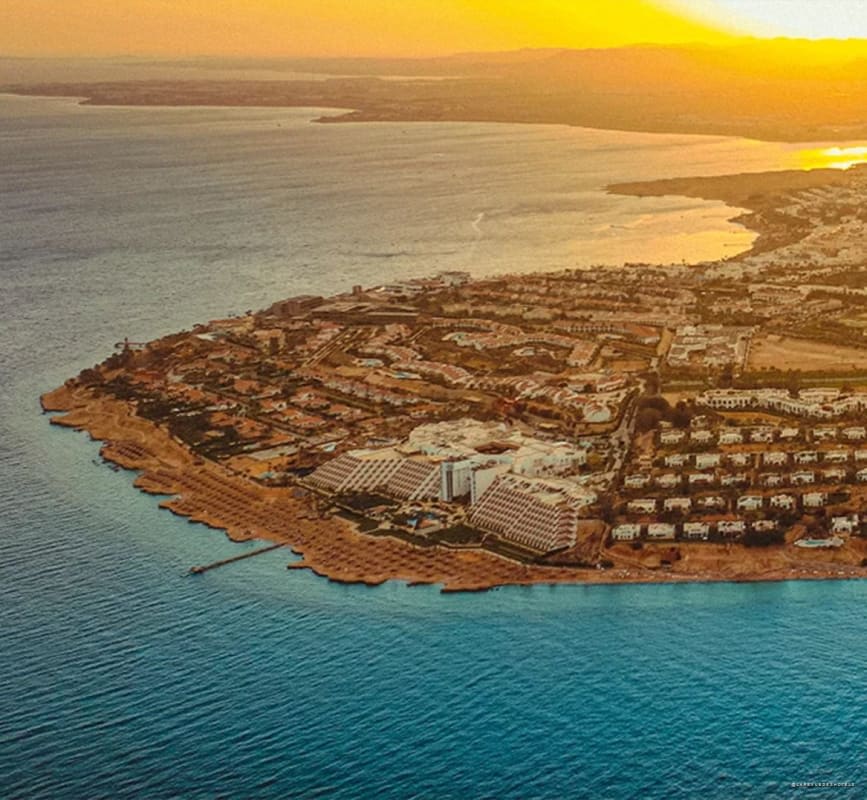 Aerial view of a coastal resort at sunset with white villas along a curved sandy shoreline and turquoise water nearby.