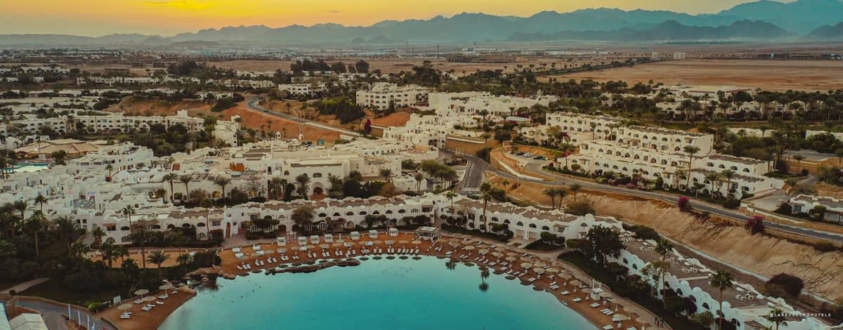 Aerial view of a coastal resort at sunset with white villas along a curved sandy shoreline and turquoise water nearby.
