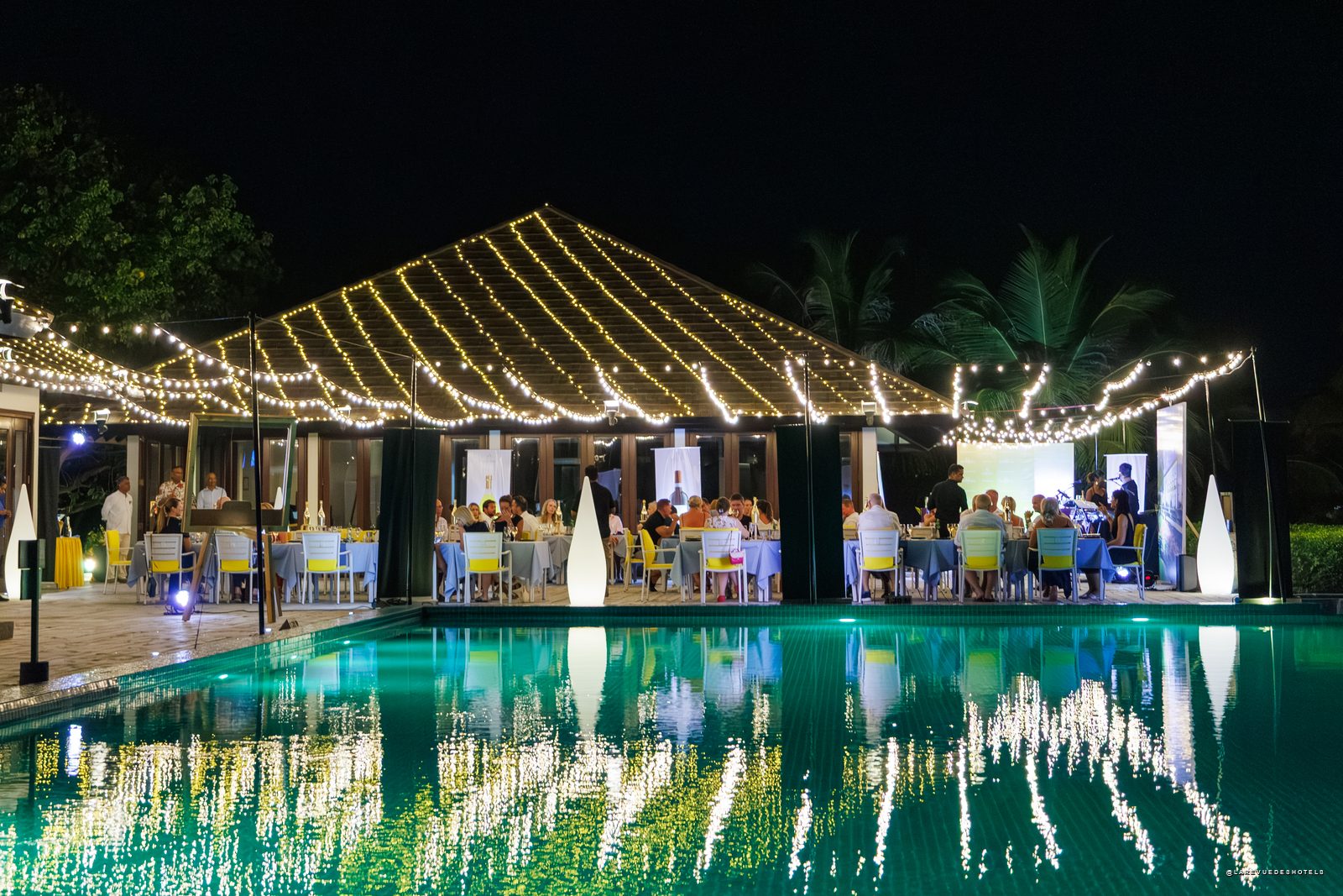 Champagne bottle centerpiece on a table with desserts and wine glasses, overlooking turquoise sea and boats in the distance.