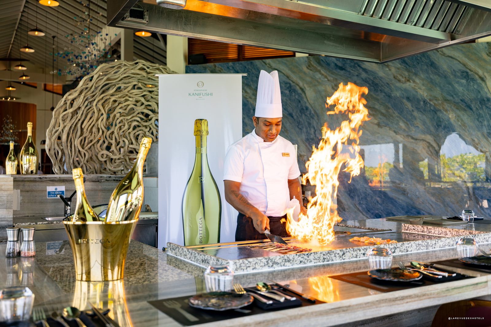 Champagne bottle centerpiece on a table with desserts and wine glasses, overlooking turquoise sea and boats in the distance.