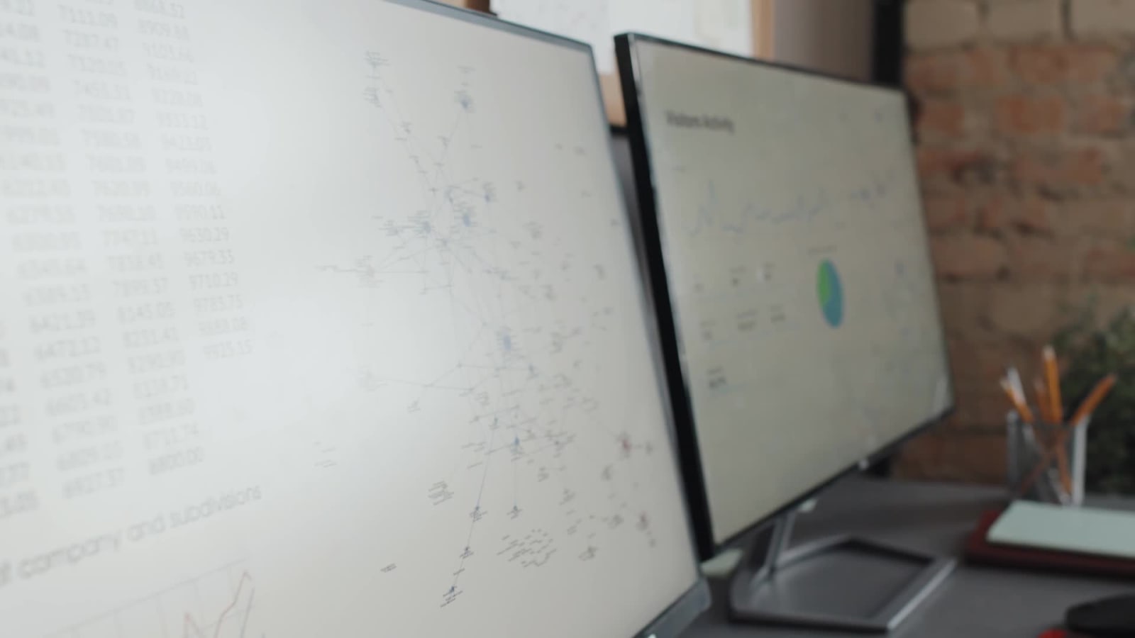 Zwei Bildschirme mit Tabellen, Netzwerkgrafik und Diagrammen in einem Büro.