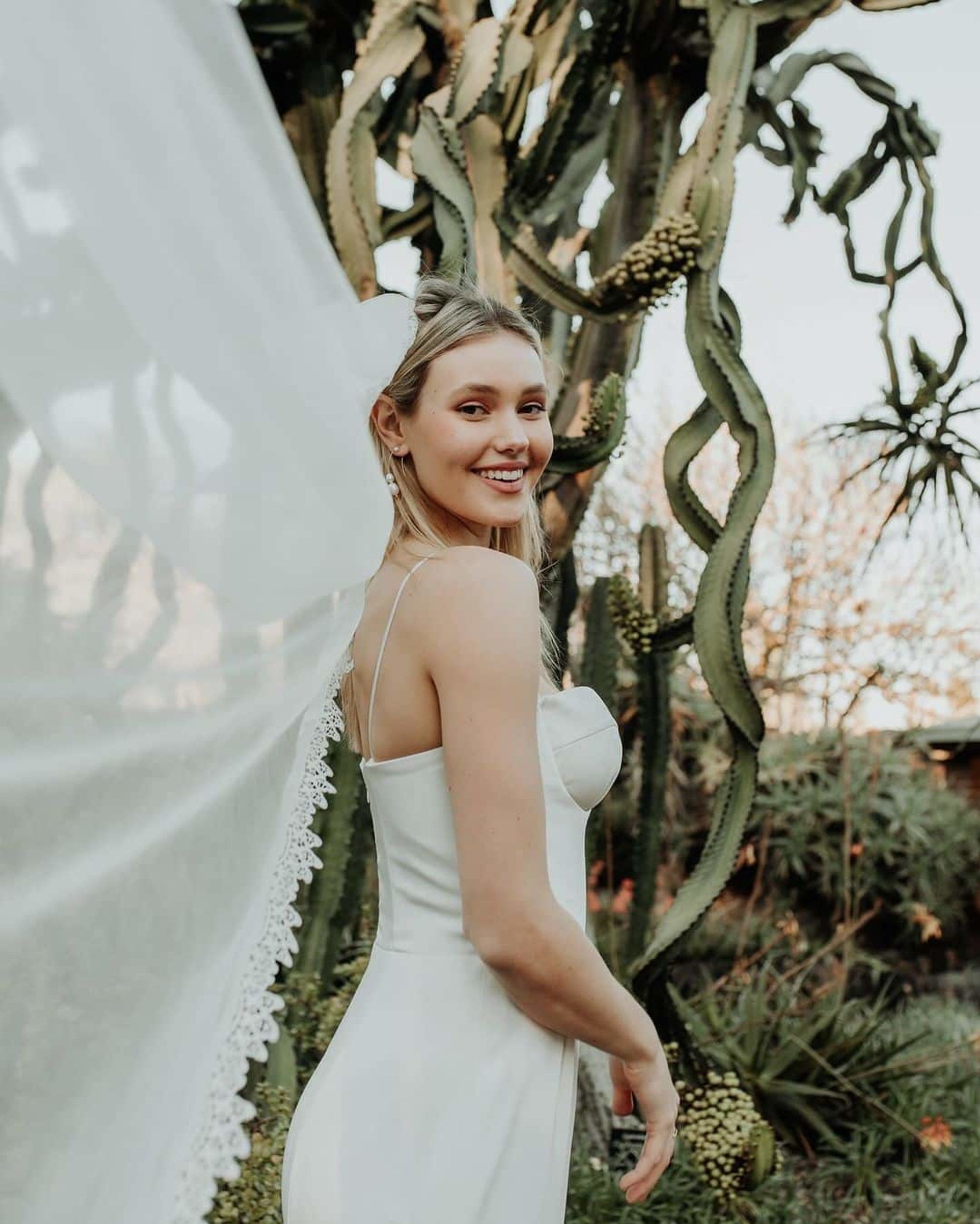 Smiling bride in a simple white gown and veil posing in a cactus garden at an outdoor wedding.