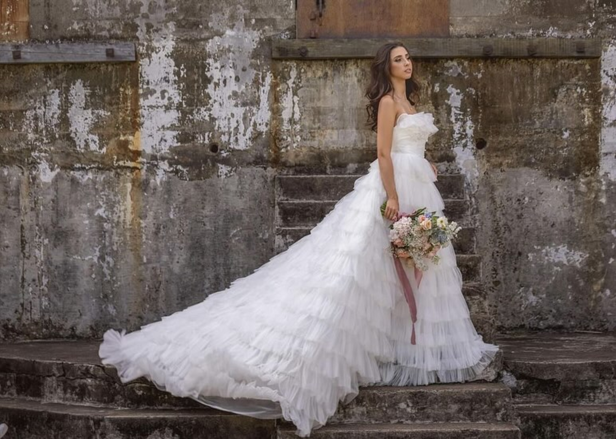 Bride in a flowing white gown holding a bouquet while standing on rustic stone steps against a weathered wall.