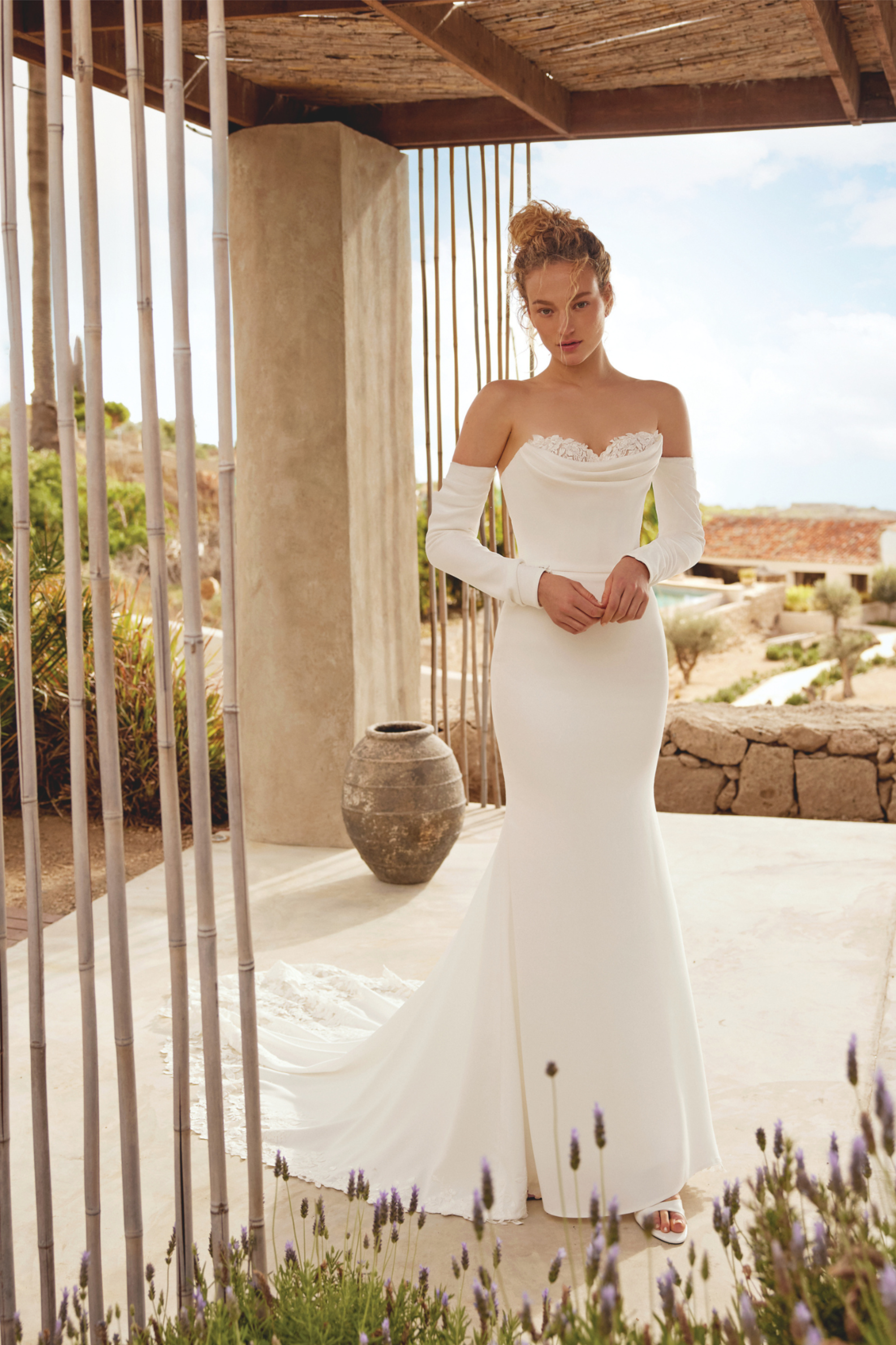 Bride in a fitted off-the-shoulder wedding dress with train standing in a rustic outdoor terrace.
