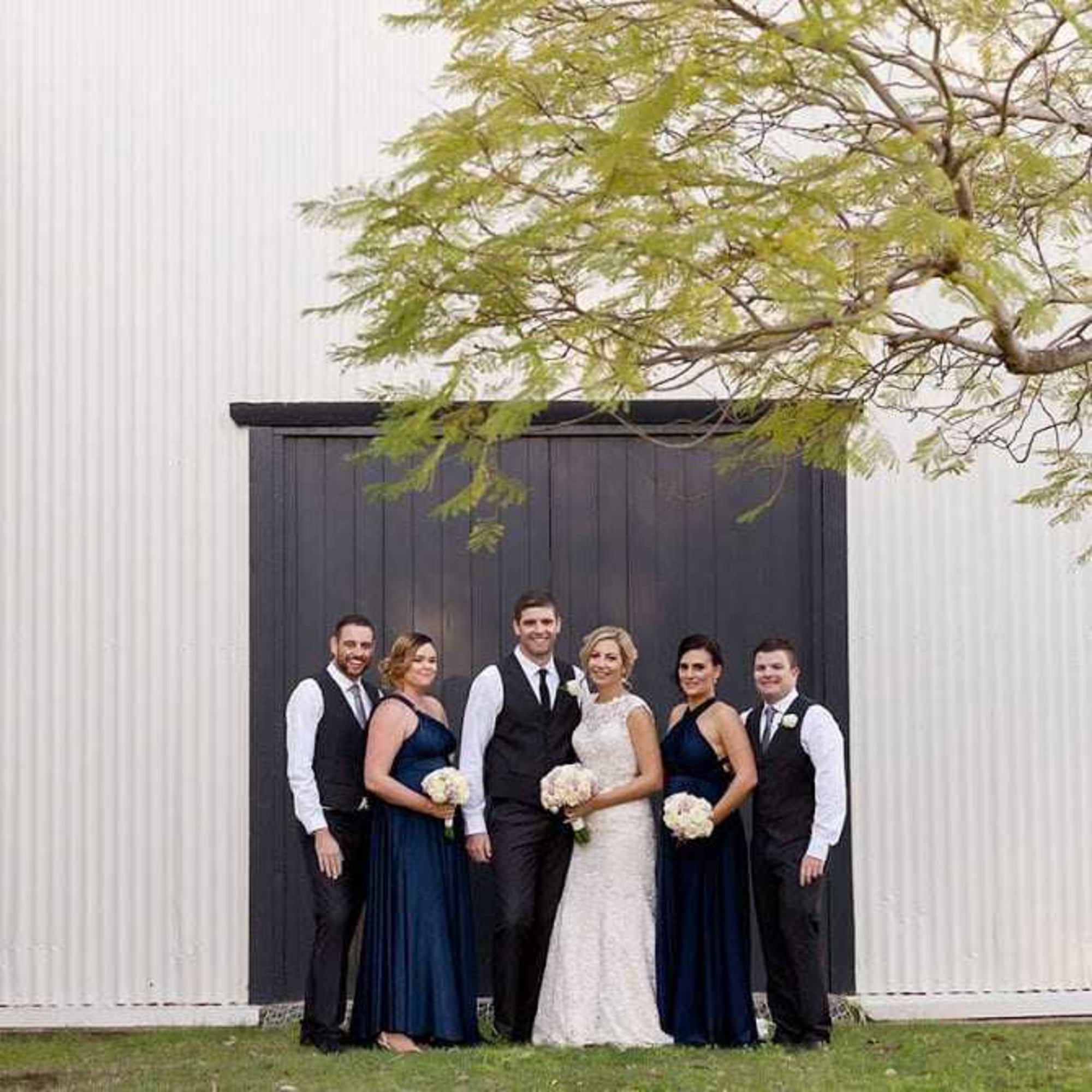 Bride and groom pose with bridesmaids and groomsmen in navy attire in front of a rustic barn door under a leafy tree.