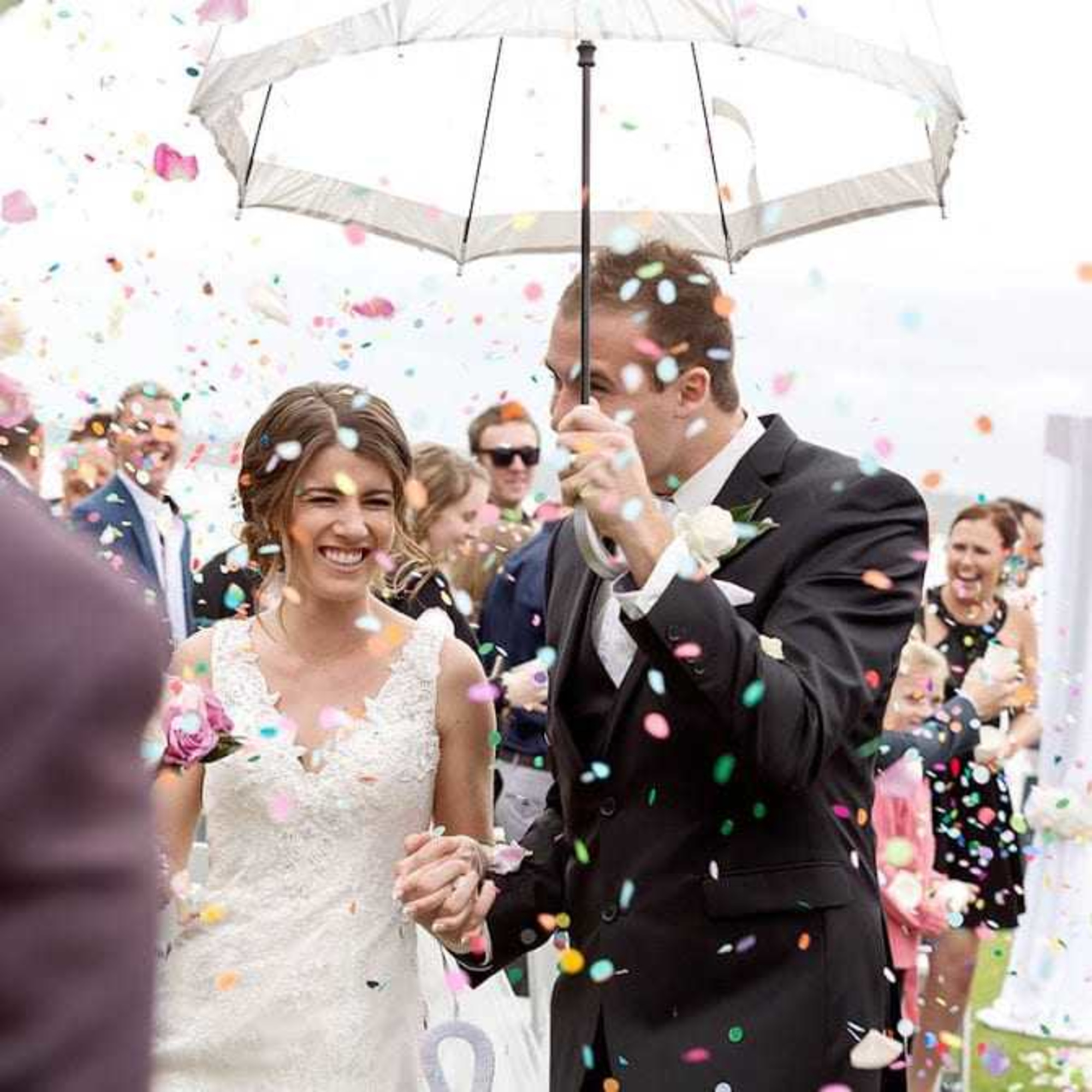 Bride and groom walk under an umbrella through colorful confetti as guests cheer during their outdoor wedding exit.