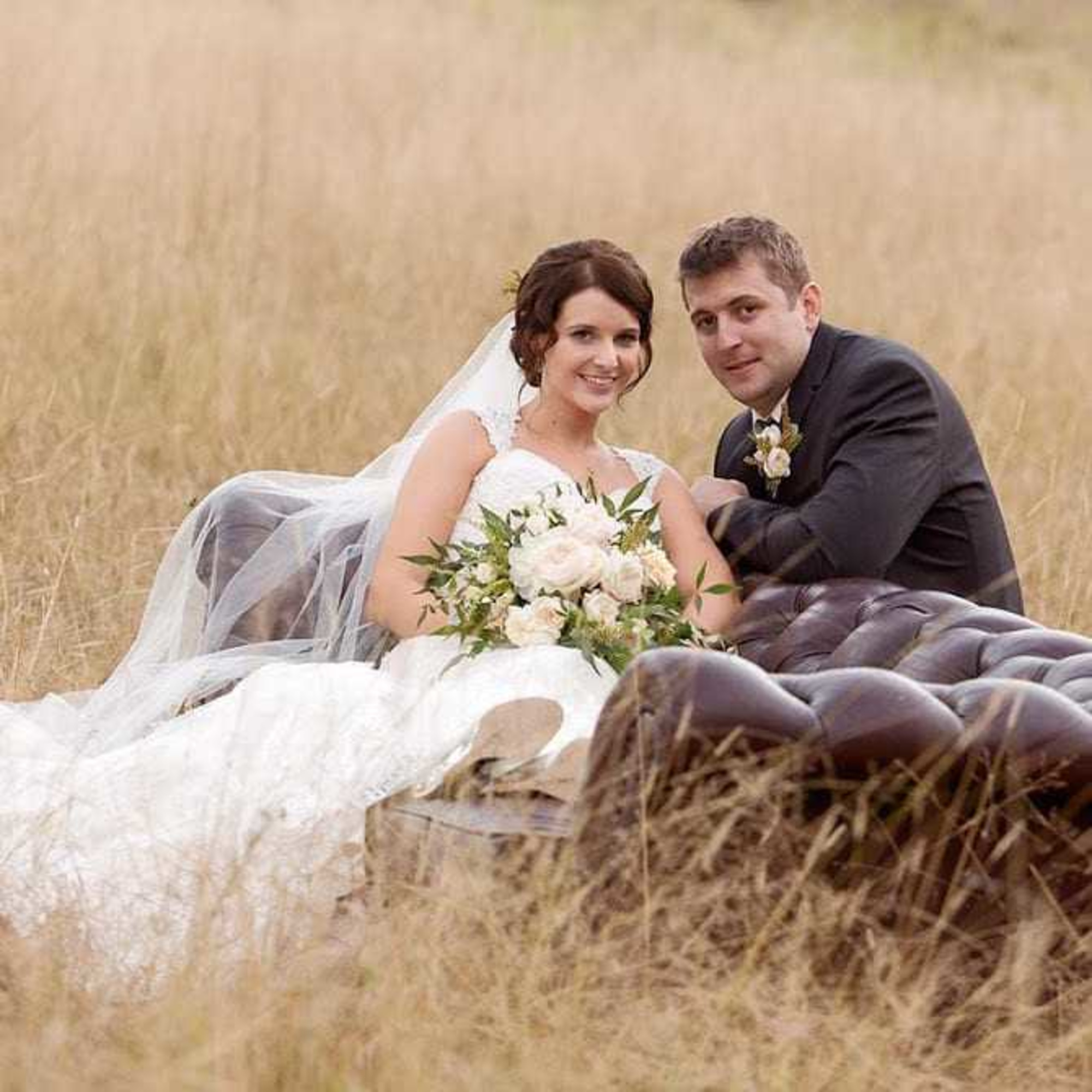 Bride and groom relax on a vintage sofa in a tall grassy field holding a soft blush and greenery bouquet.