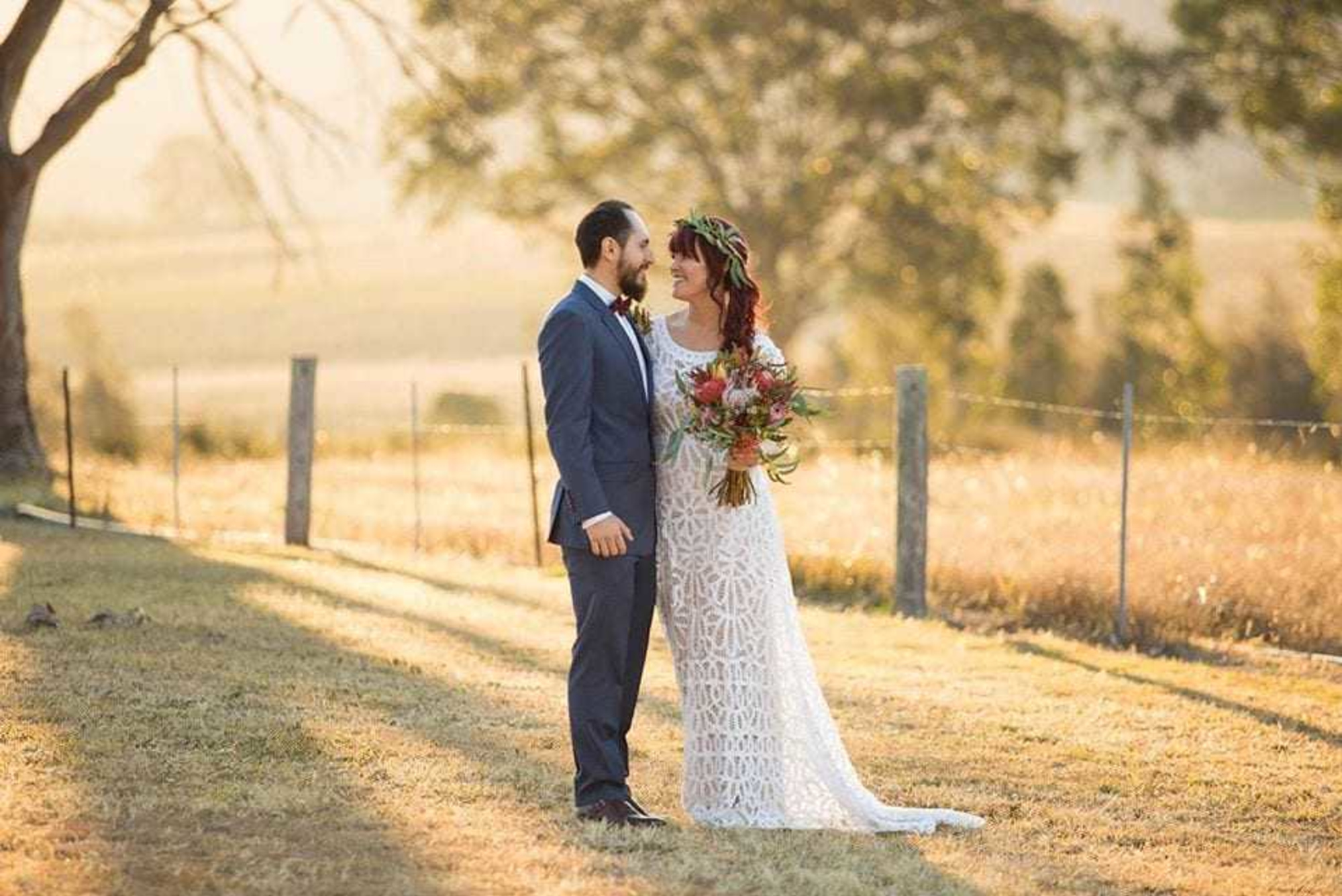 Bride and groom standing together in a sunlit rustic field holding a colorful bouquet.