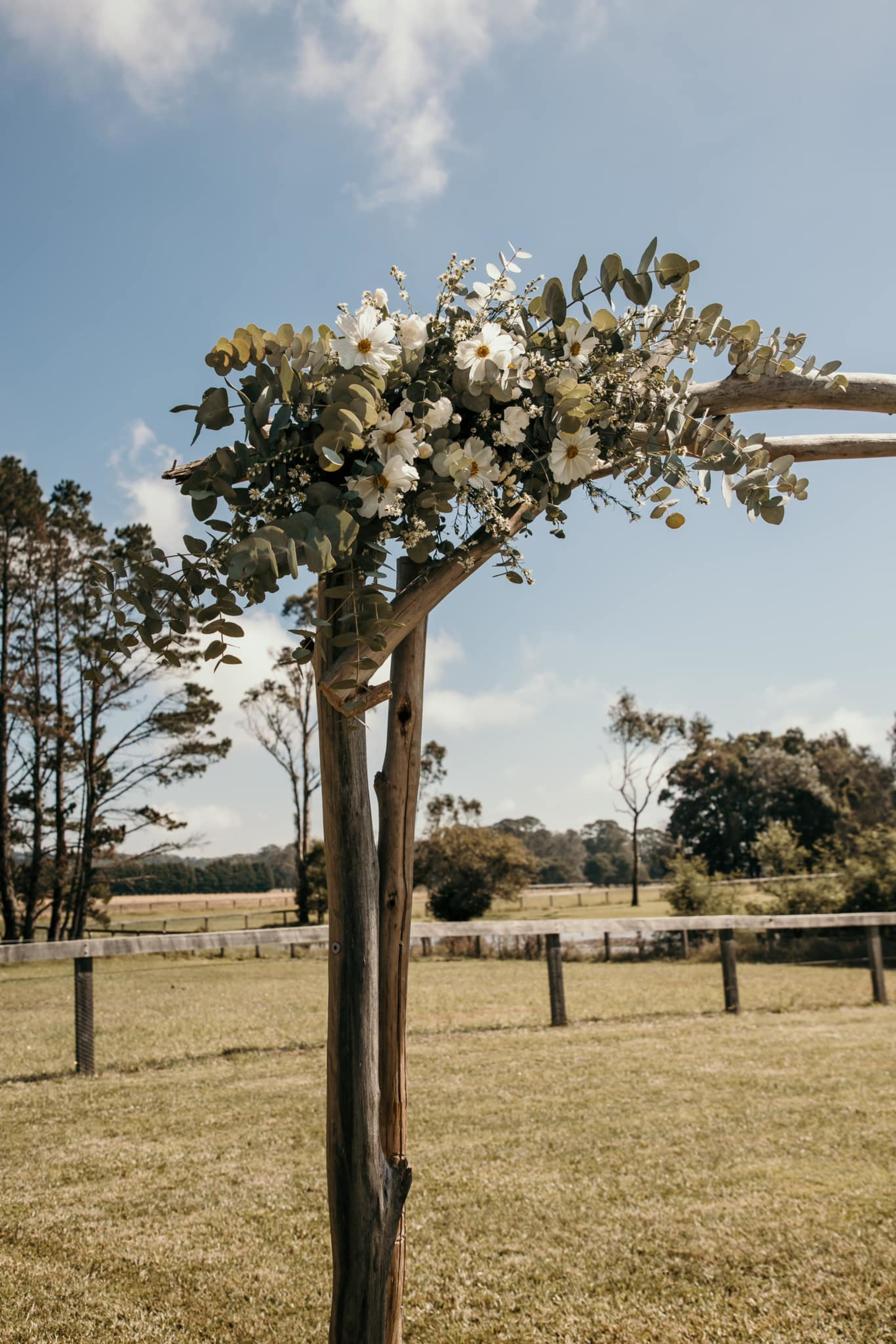 Rustic wooden wedding arch decorated with white flowers and greenery in an open countryside field.