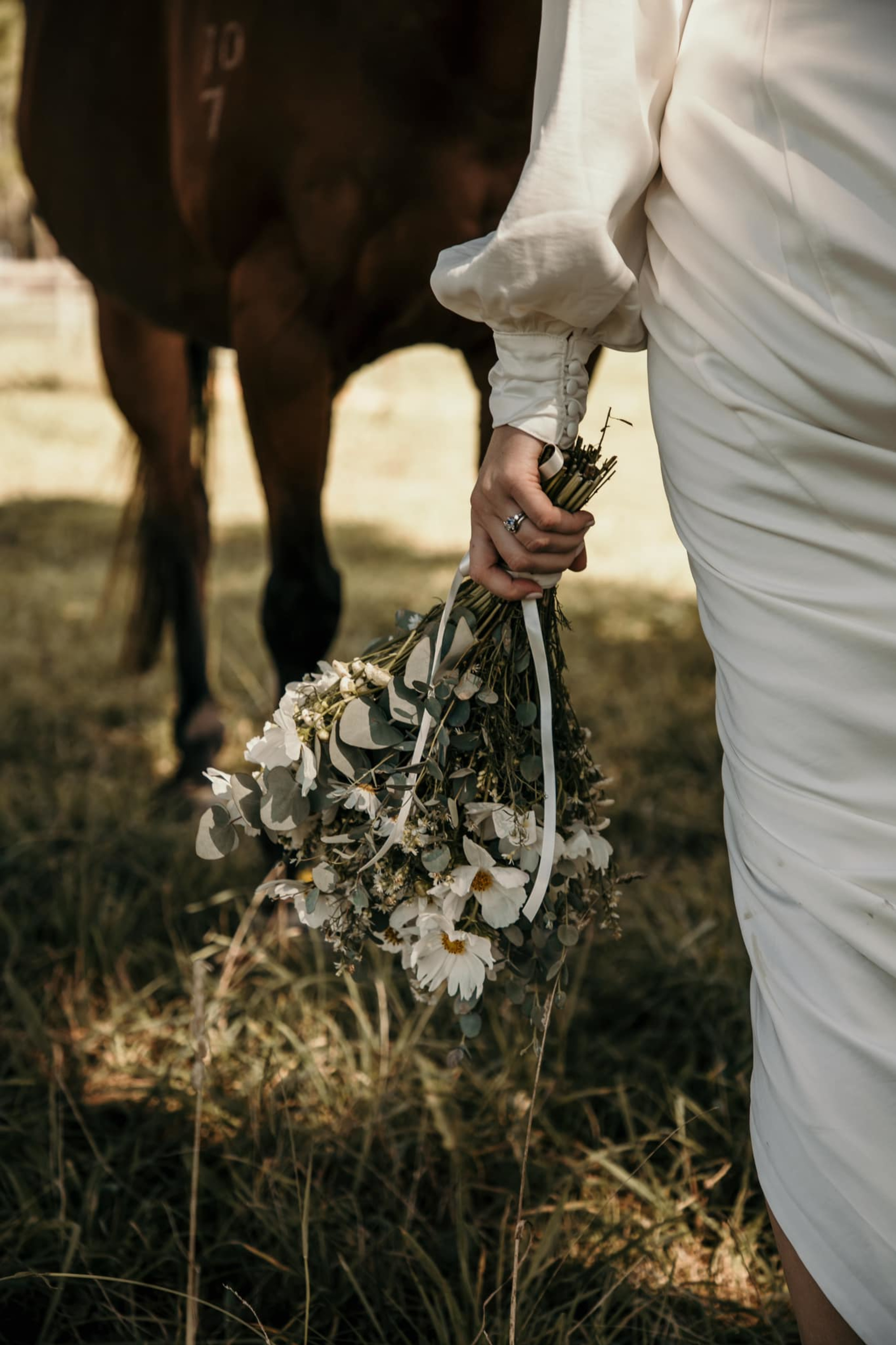 Bride in a long-sleeve gown holds a wildflower bouquet while standing beside a horse in a grassy field.