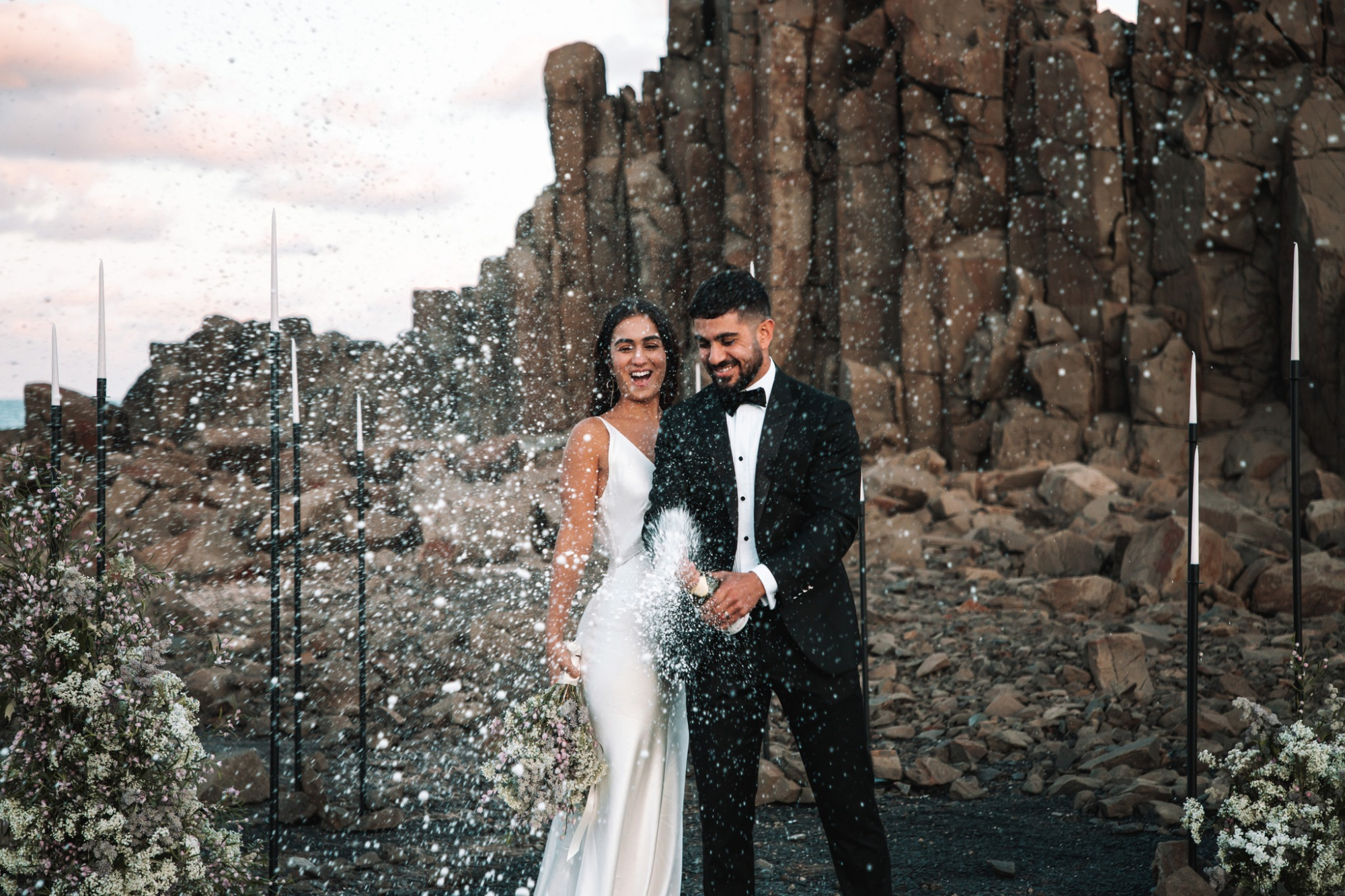 Joyful couple popping champagne in front of dramatic rocky cliffs at their outdoor wedding ceremony.