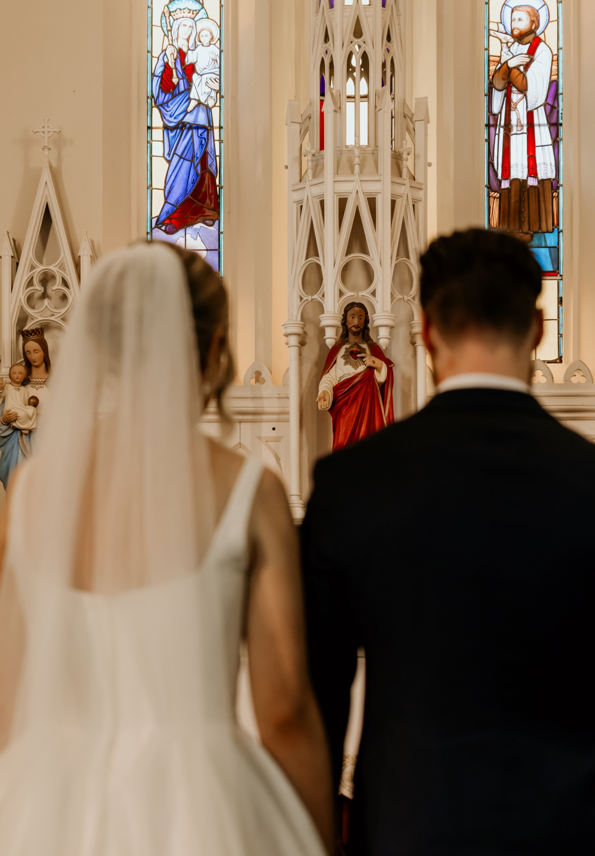 Bride and groom stand before a church altar with religious statues and stained glass windows during their wedding ceremony.