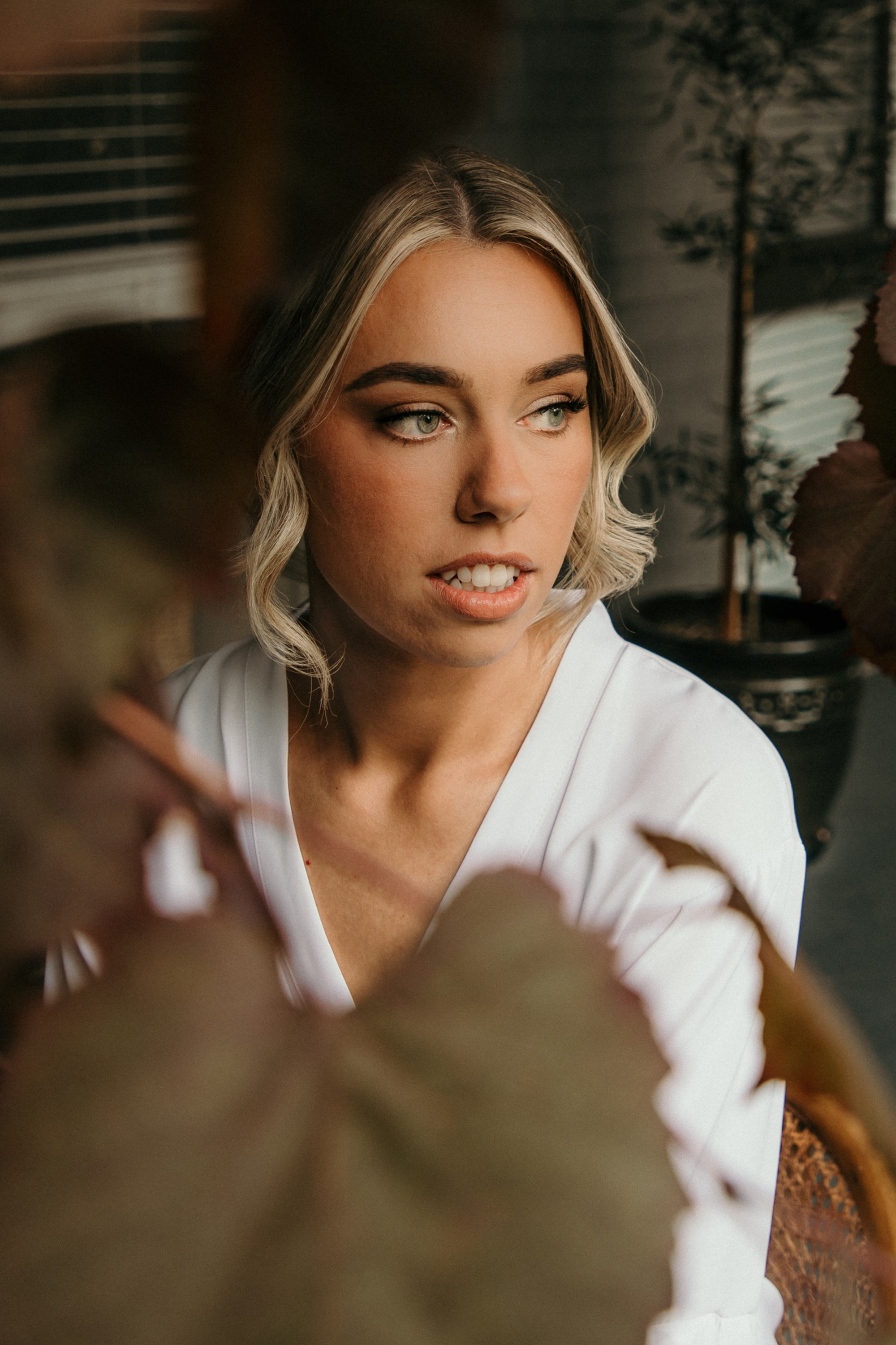 Soft, intimate bridal portrait of a woman in a white robe framed by blurred foliage indoors.