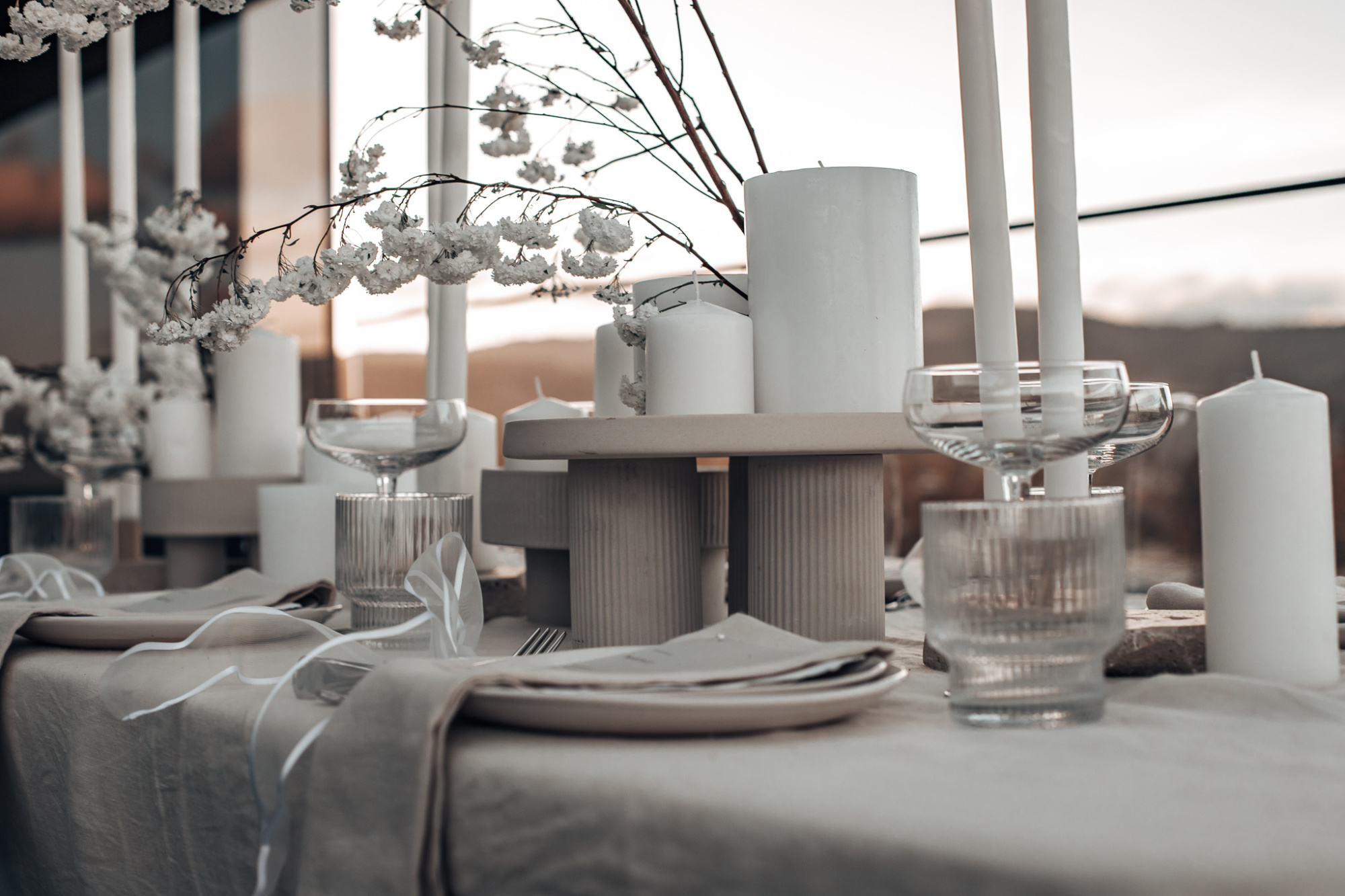 Minimalist outdoor wedding tablescape with neutral linens, white candles, glassware, and delicate white florals at sunset.