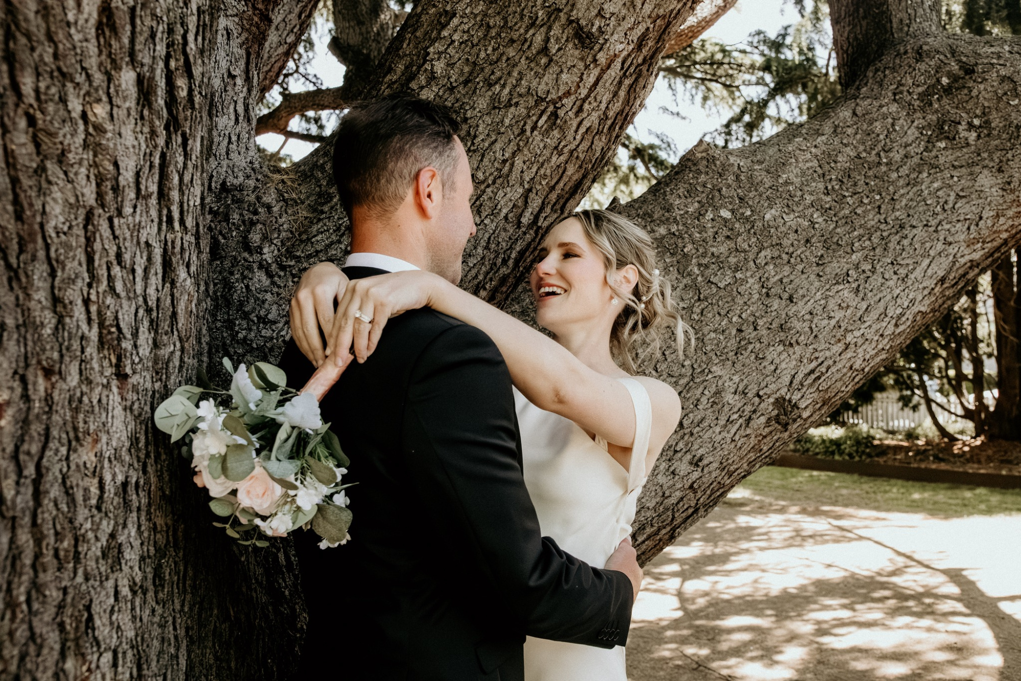 Bride and groom share a joyful embrace under a large tree, with the bride holding a soft pastel bouquet.