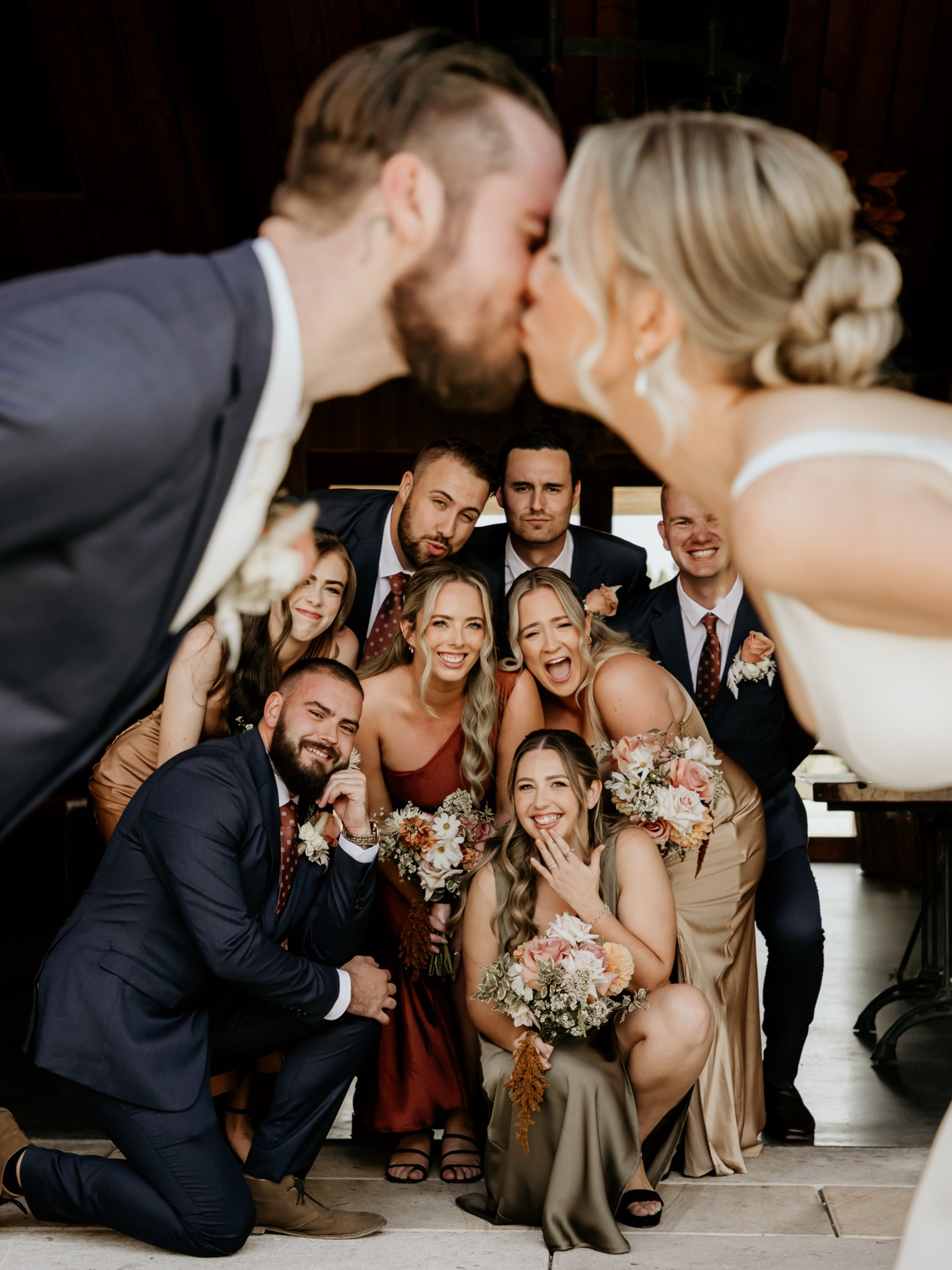 Joyful wedding party poses behind the bride and groom kissing in a candid group photo.