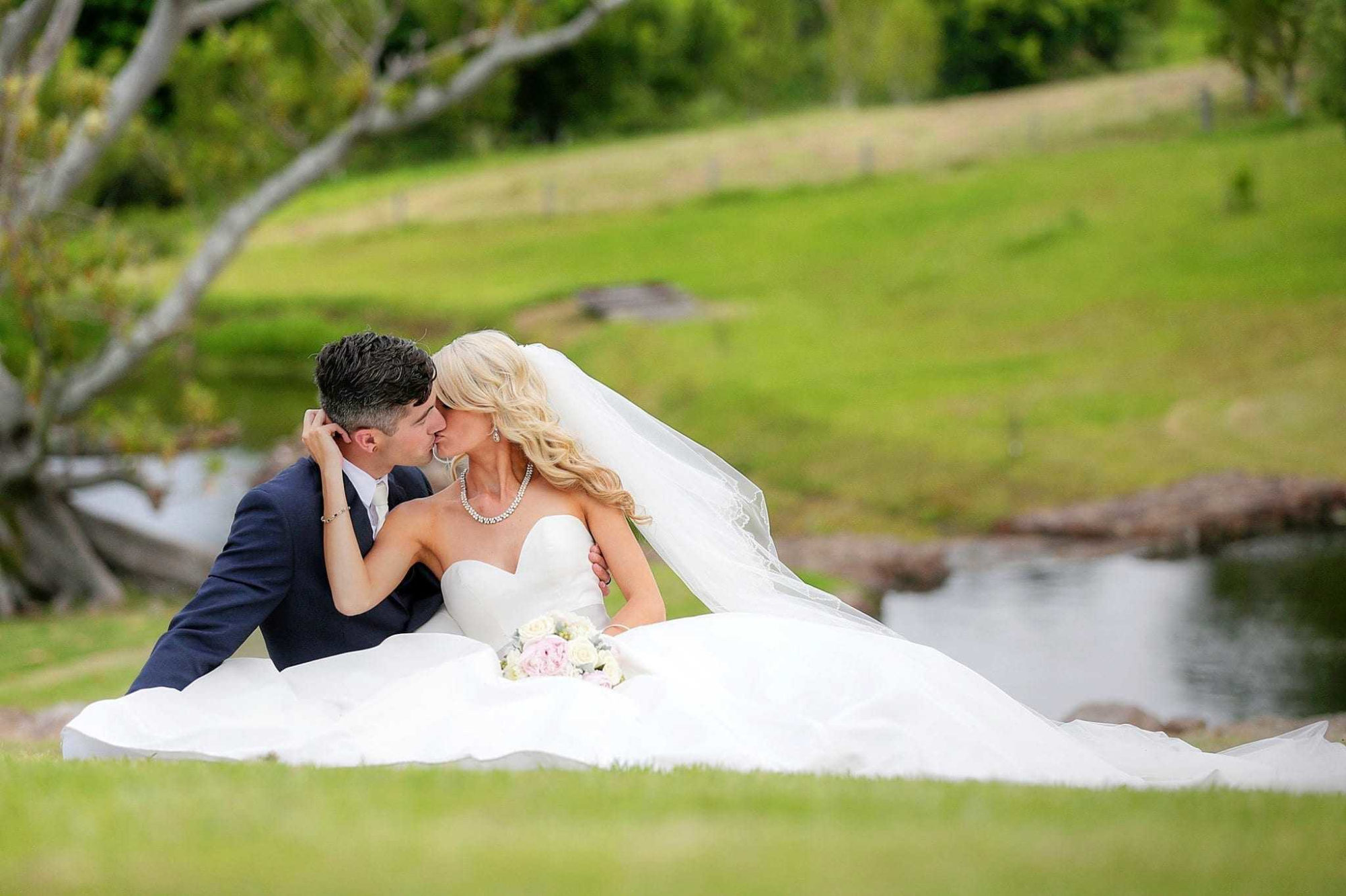 Bride and groom share a kiss while sitting on the grass by a pond in a lush outdoor setting.