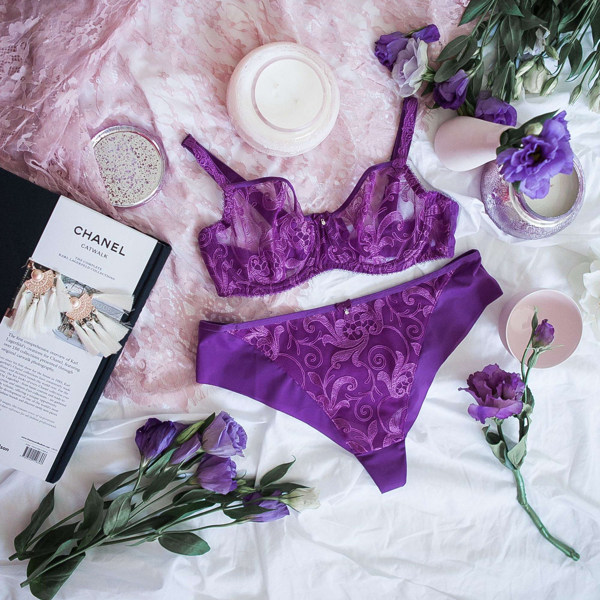 Flat lay of purple lace bridal lingerie with flowers, candles, and a fashion book on soft pink and white fabric.
