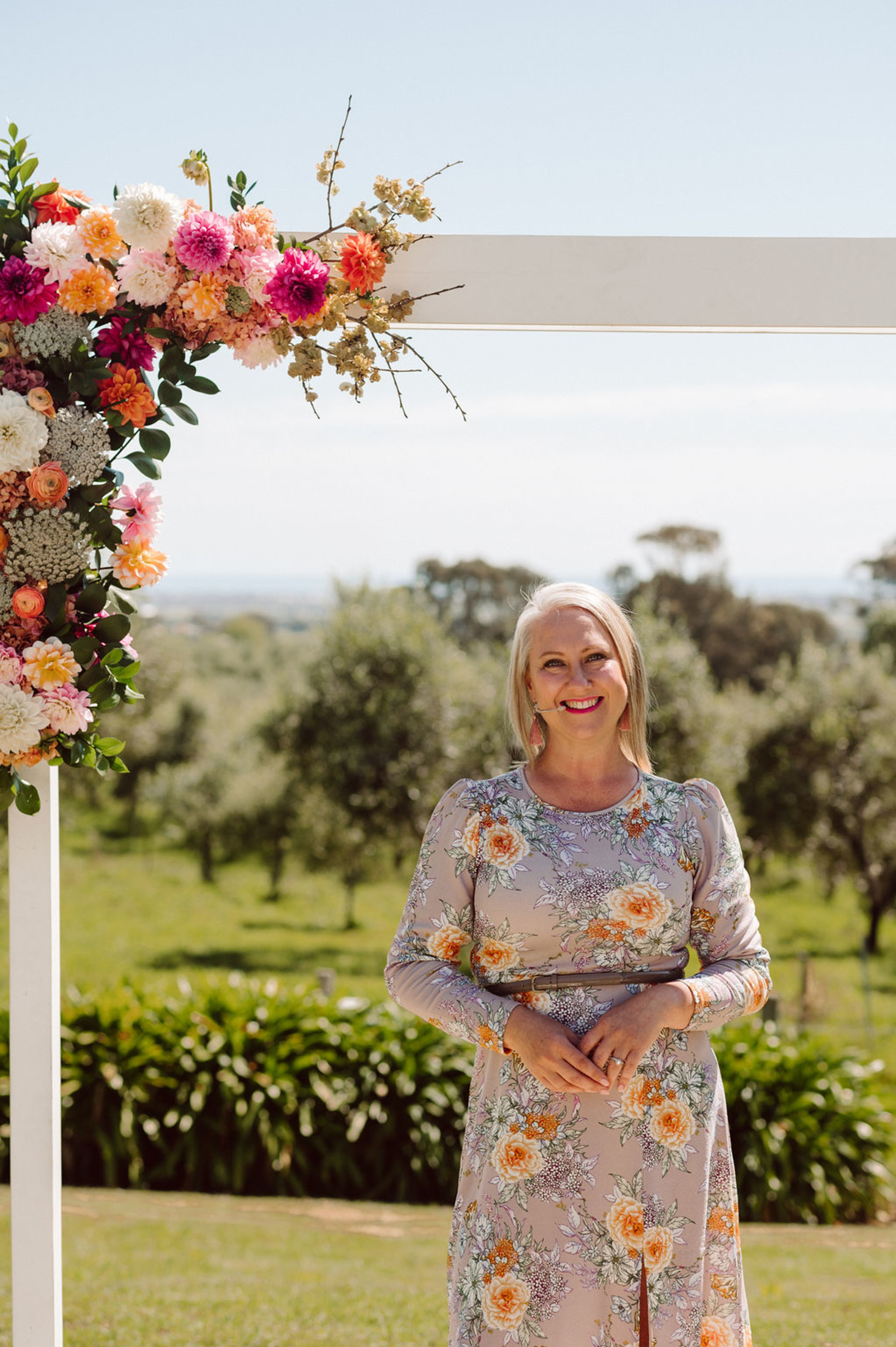 A smiling woman in a floral dress stands by a beautiful flower-adorned arch, perfect for your outdoor wedding ceremony.