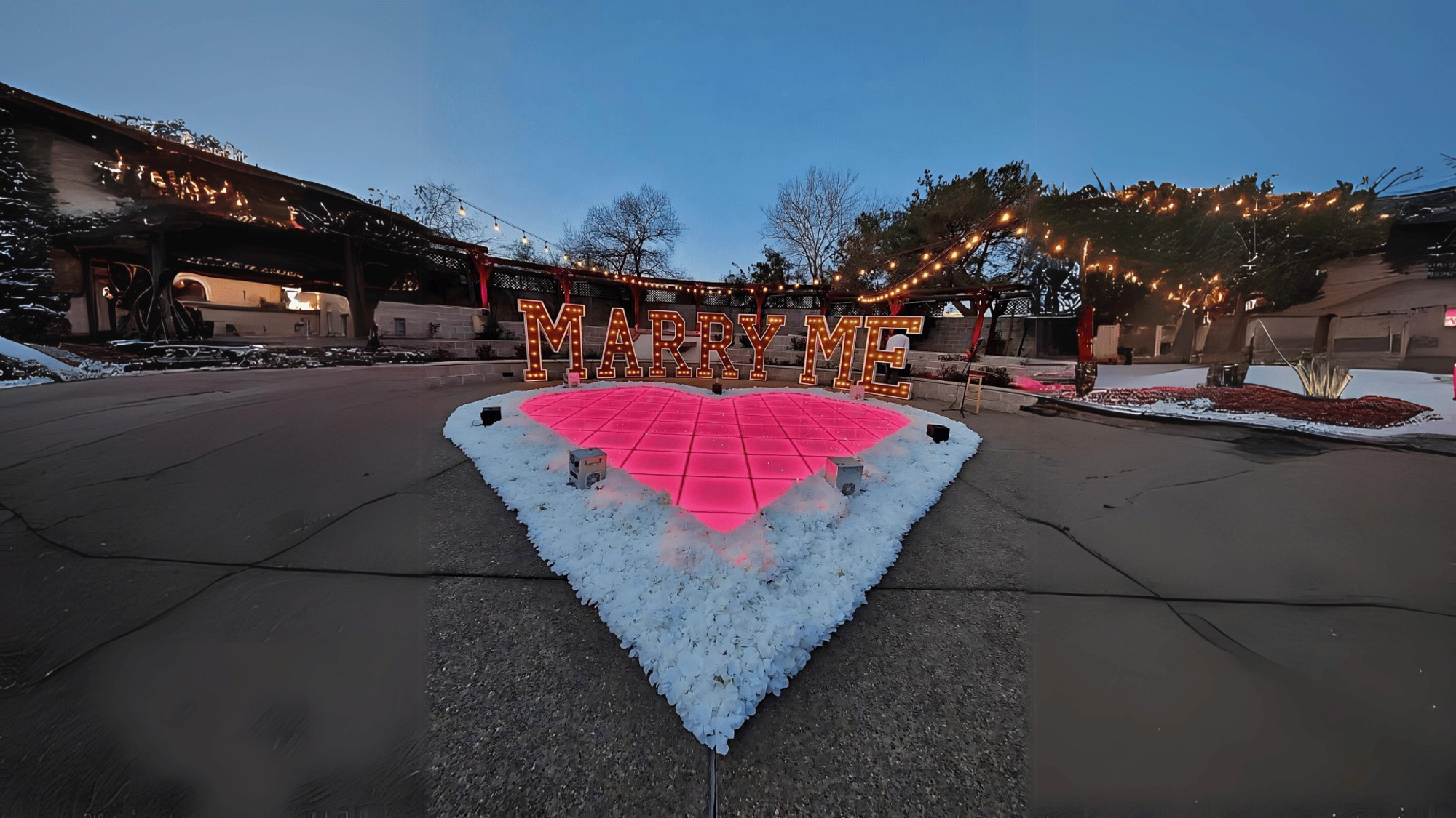 Outdoor proposal setup with illuminated MARRY ME letters and a glowing pink heart surrounded by flowers and string lights.