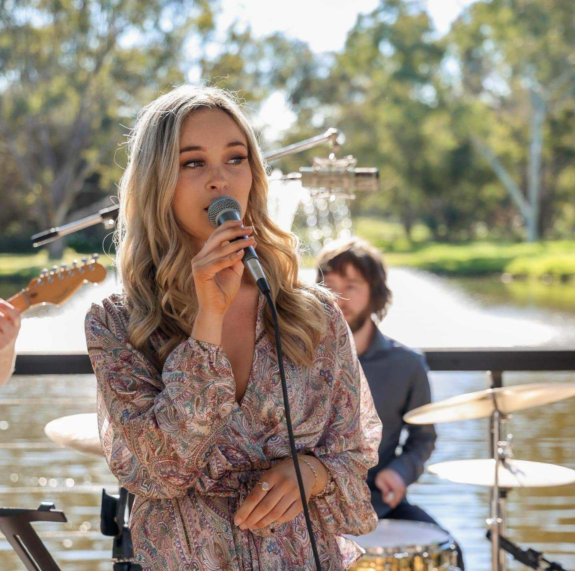 Female vocalist sings into a microphone with a live band performing by a scenic lakeside backdrop.
