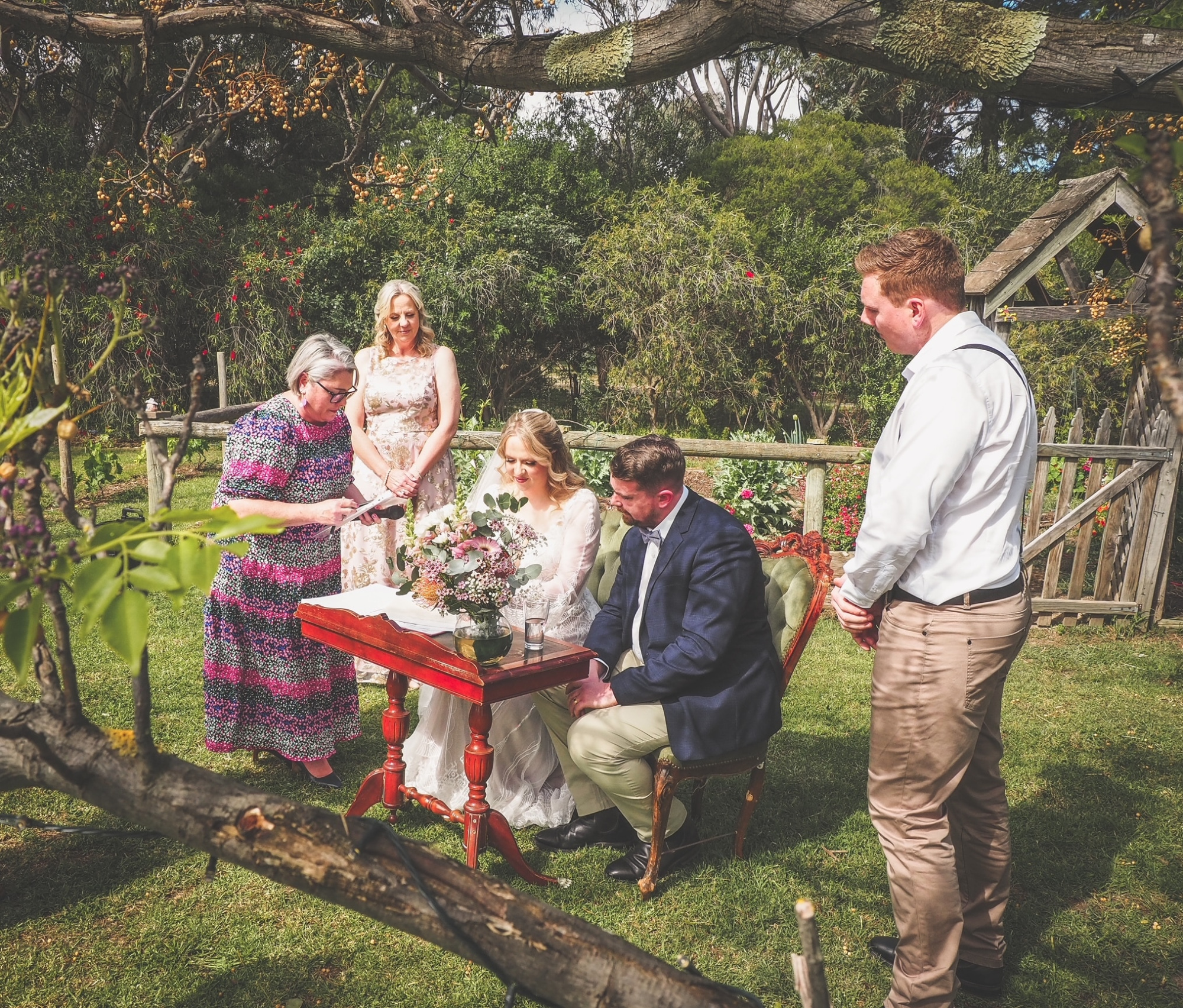 Couple signs their marriage documents at an intimate outdoor garden ceremony with celebrant and attendants.