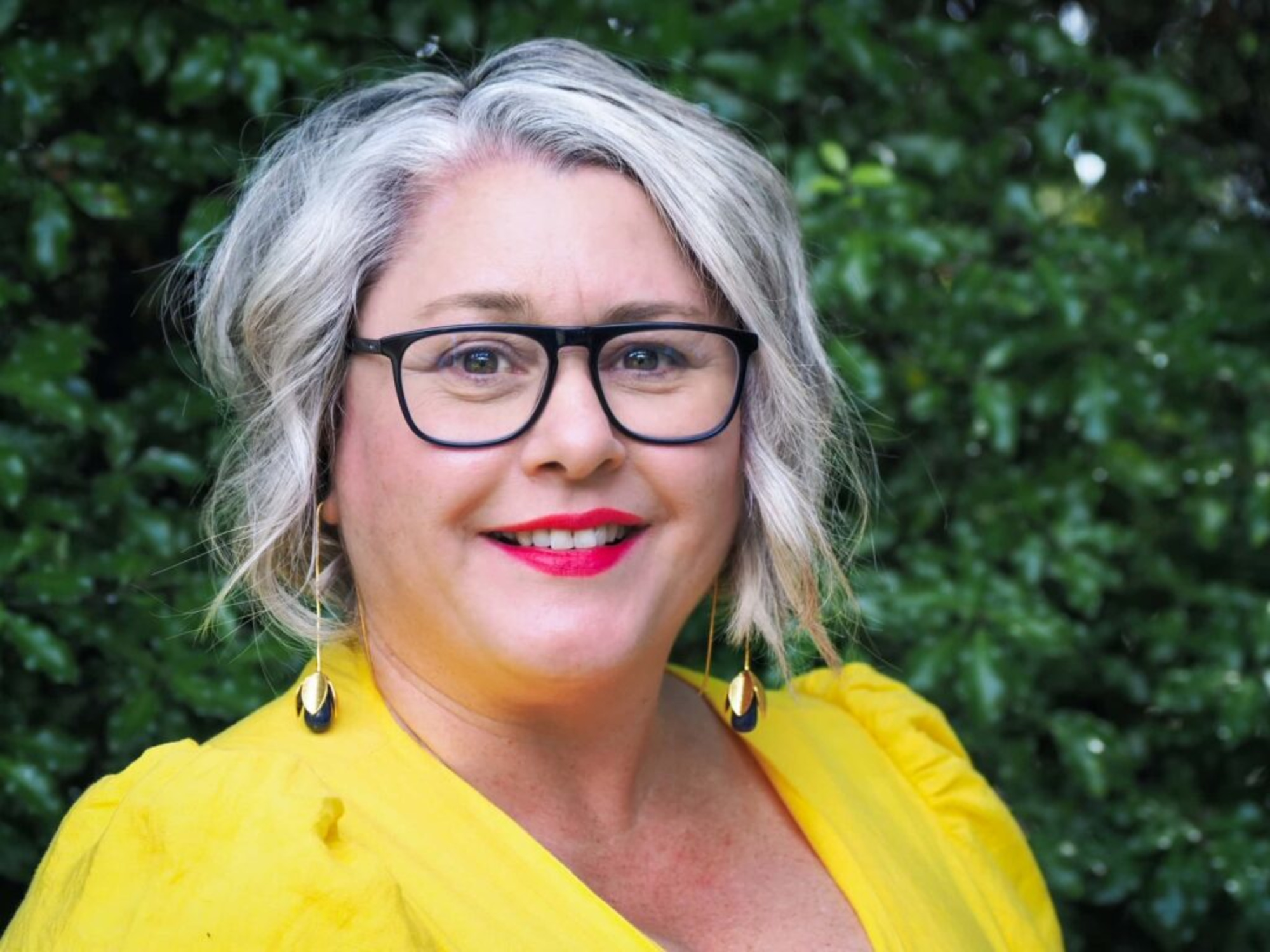 Smiling wedding celebrant in a bright yellow outfit standing outdoors in front of green foliage.