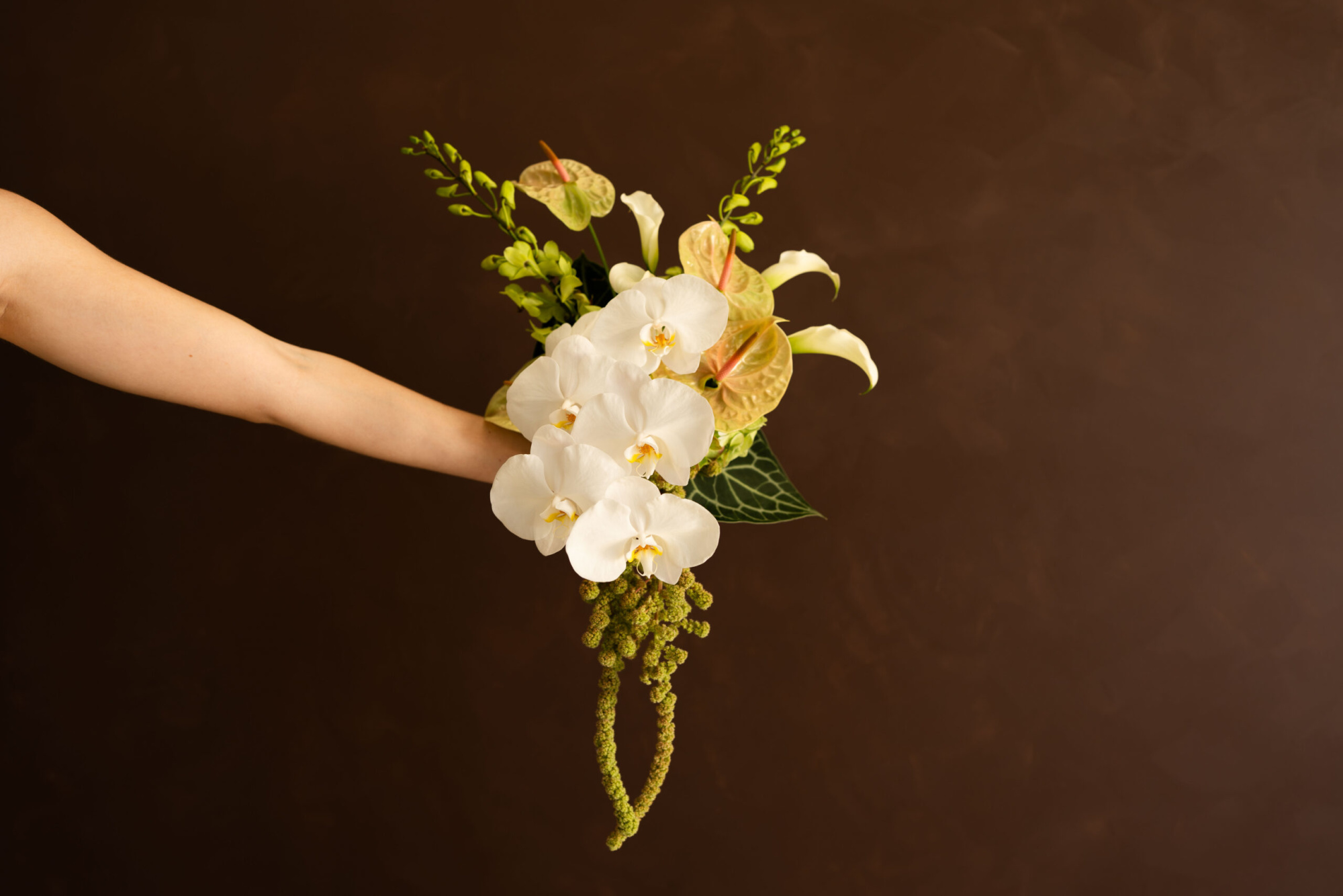 Outstretched arm holding a cascading white orchid and green tropical wedding bouquet against a brown background.