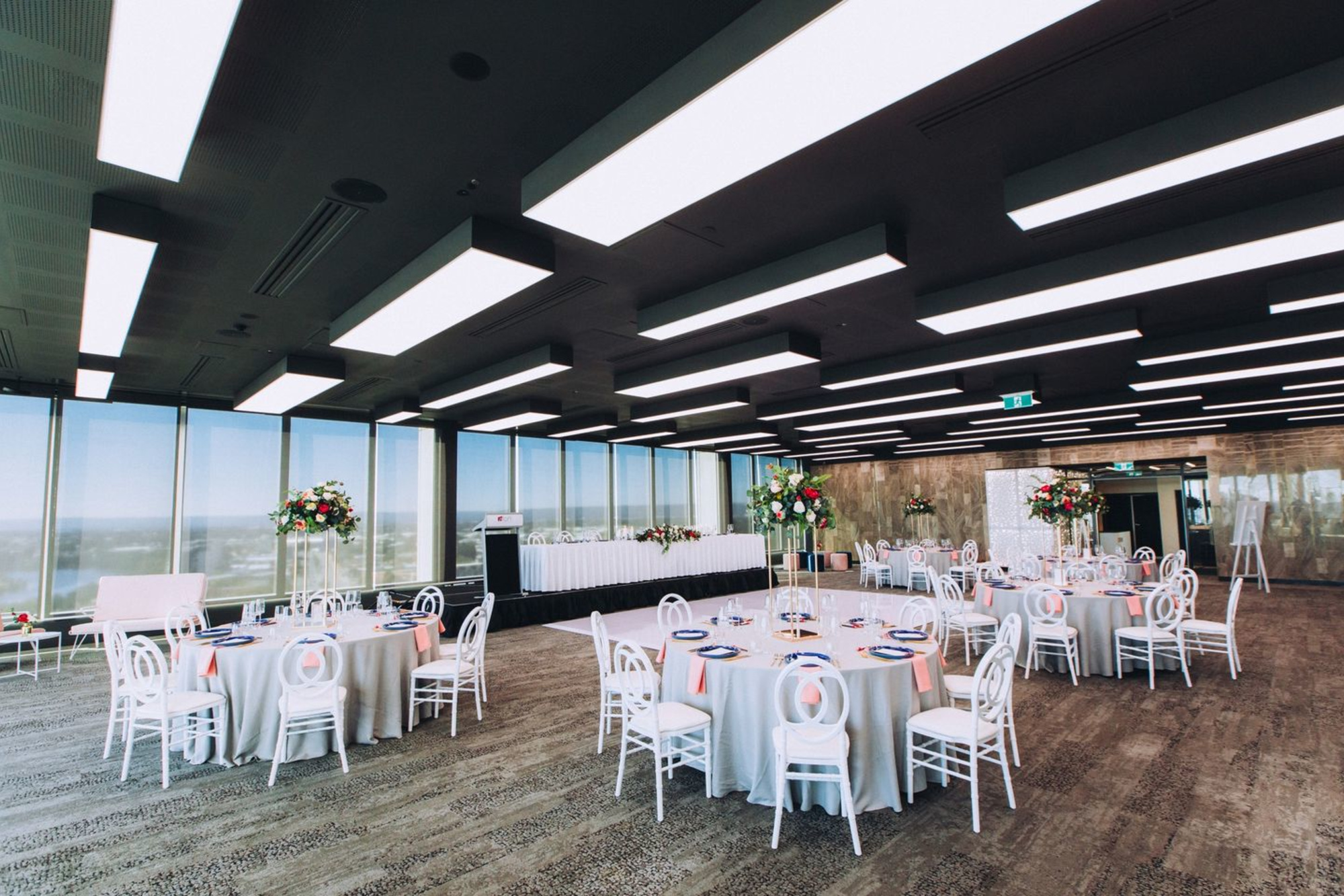 Modern indoor wedding reception venue with round tables, tall floral centerpieces, and large windows overlooking the city.