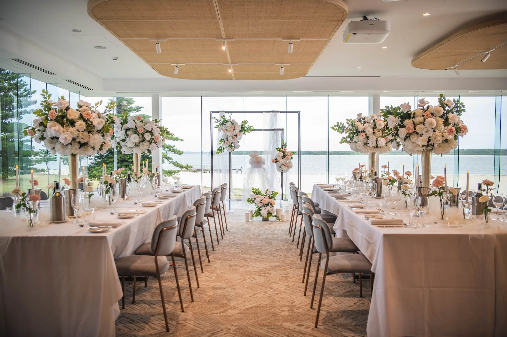 Elegant waterfront wedding reception setup with long tables, tall floral centerpieces, and large windows overlooking the water.