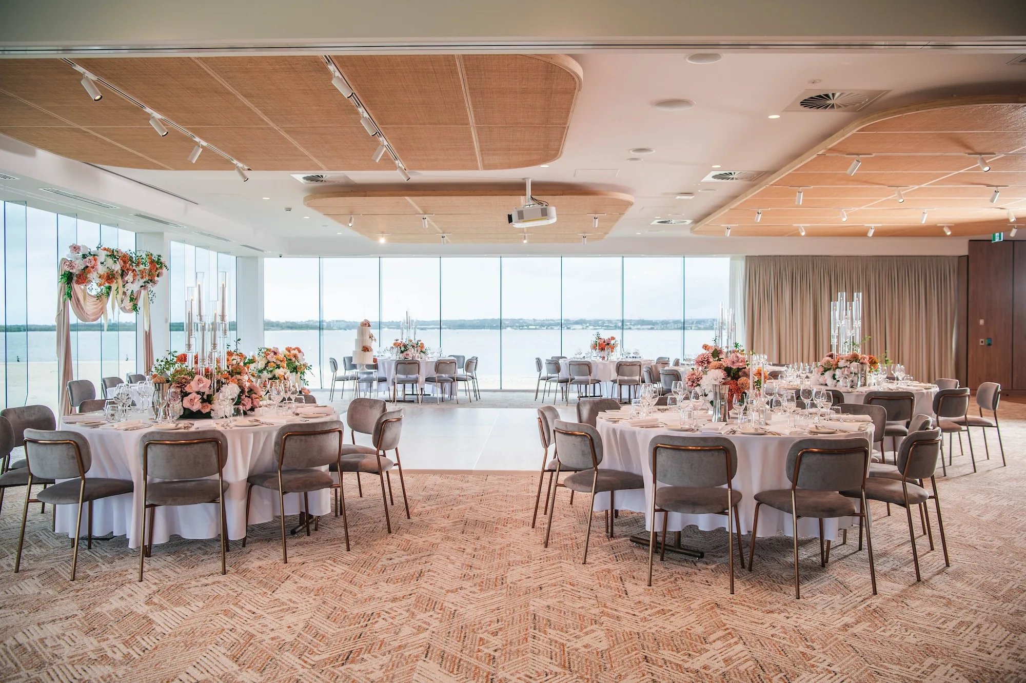 Elegant waterfront wedding reception room with round tables, floral centerpieces, and panoramic windows overlooking the water.