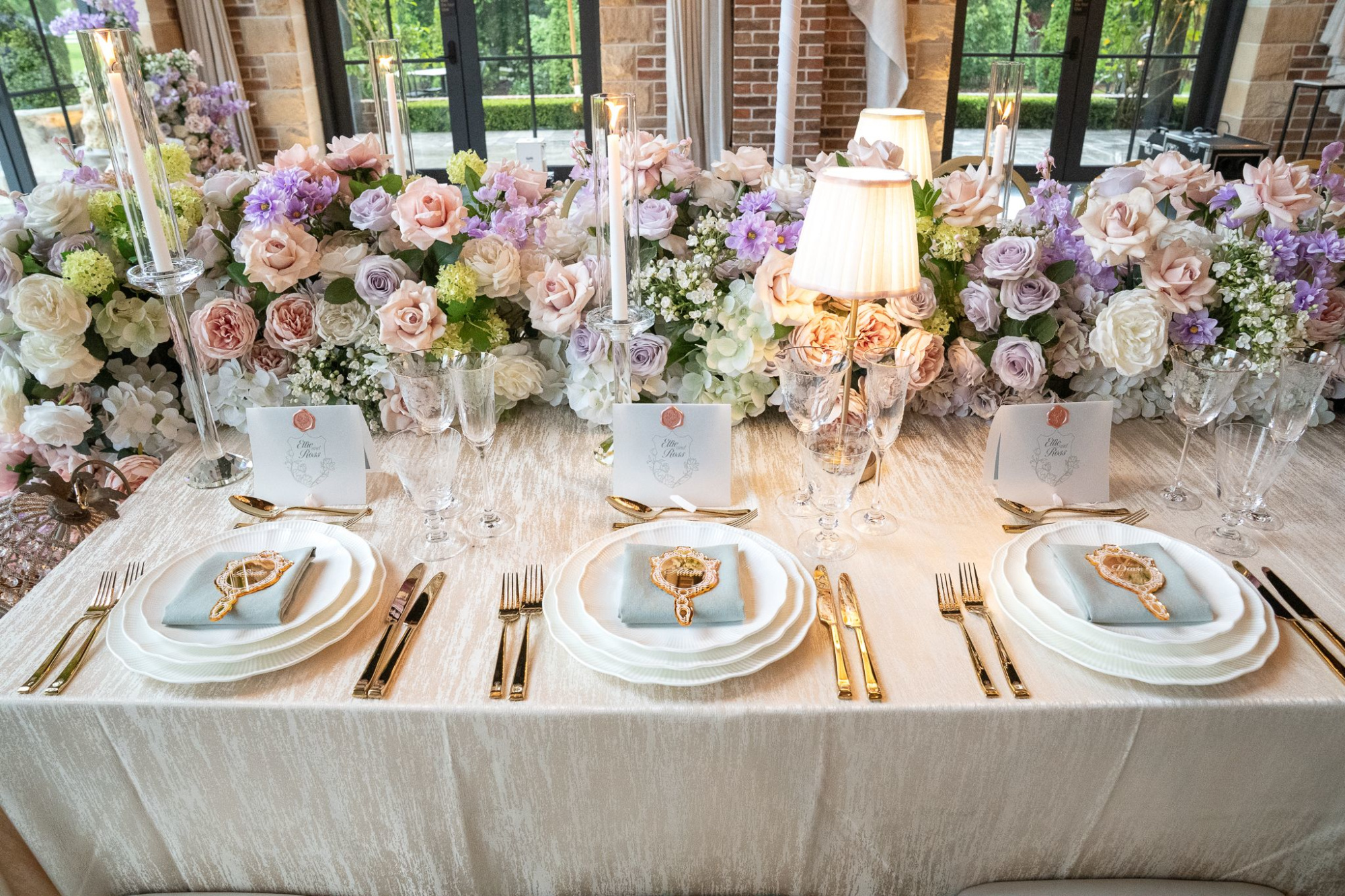 Elegant wedding reception table with pastel floral garland, gold flatware, and candlelit place settings.