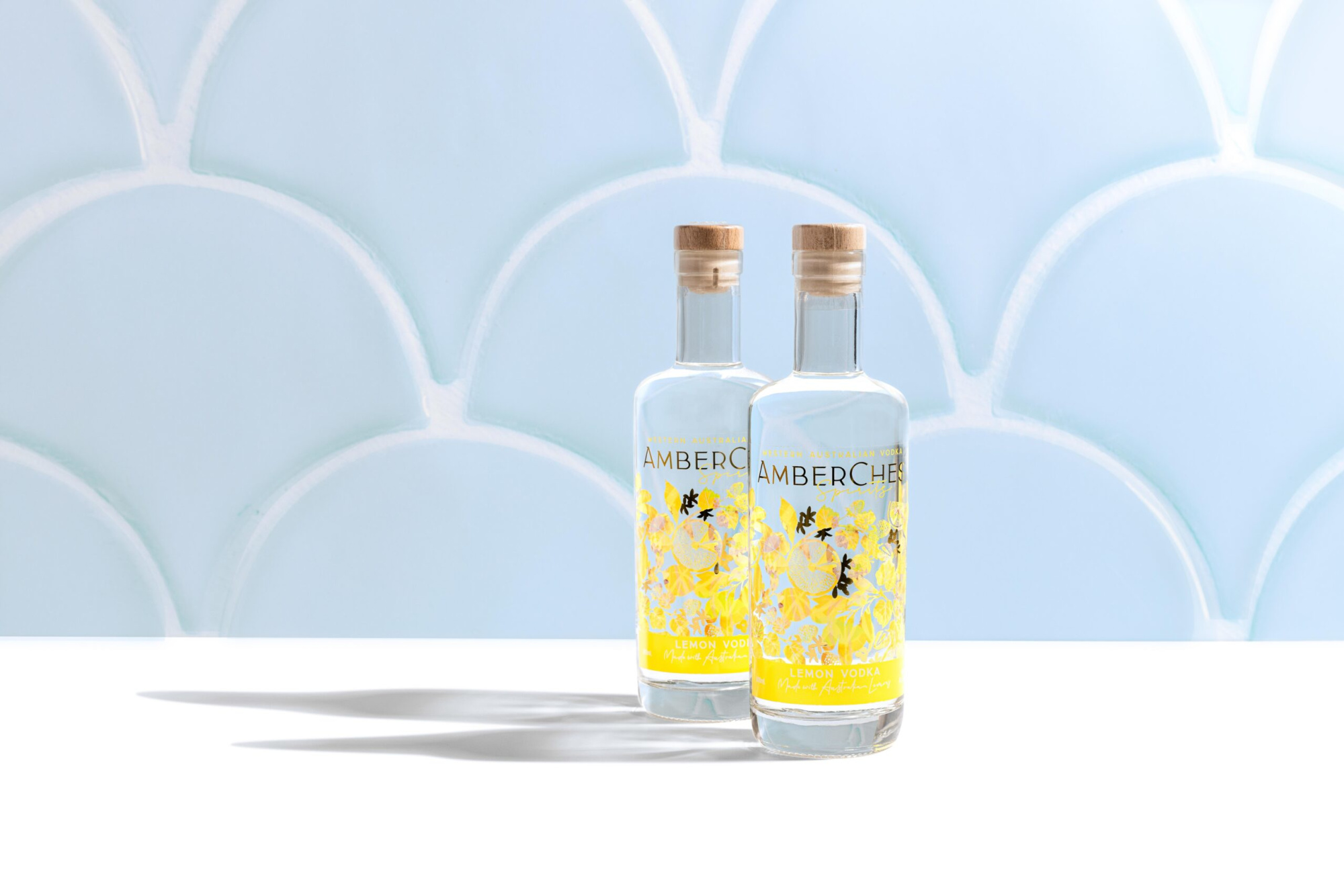Two lemon vodka bottles with floral labels stand on a white surface against a light blue patterned backdrop.
