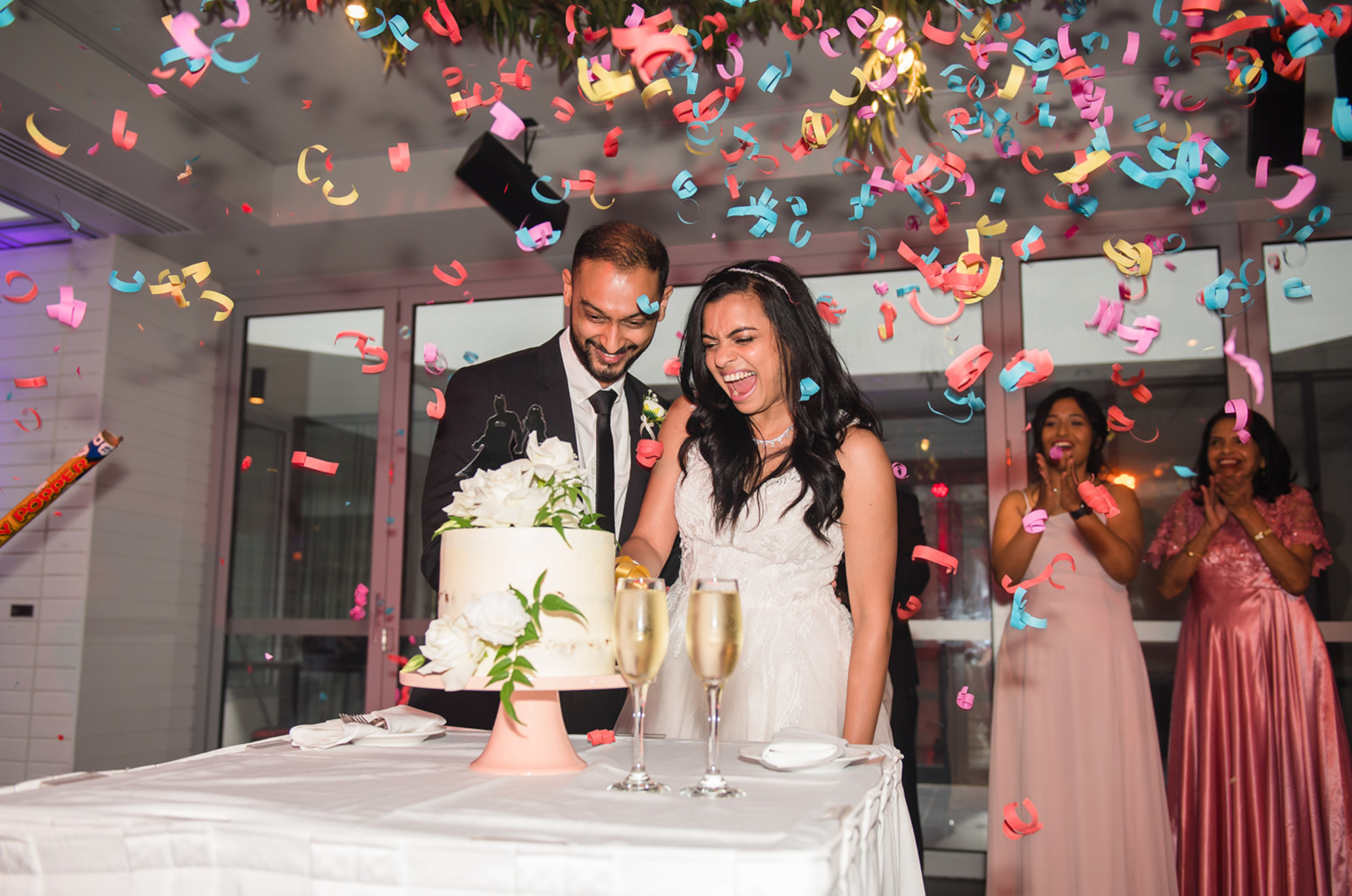 Joyful couple cuts their wedding cake as colorful confetti falls and guests cheer around them.