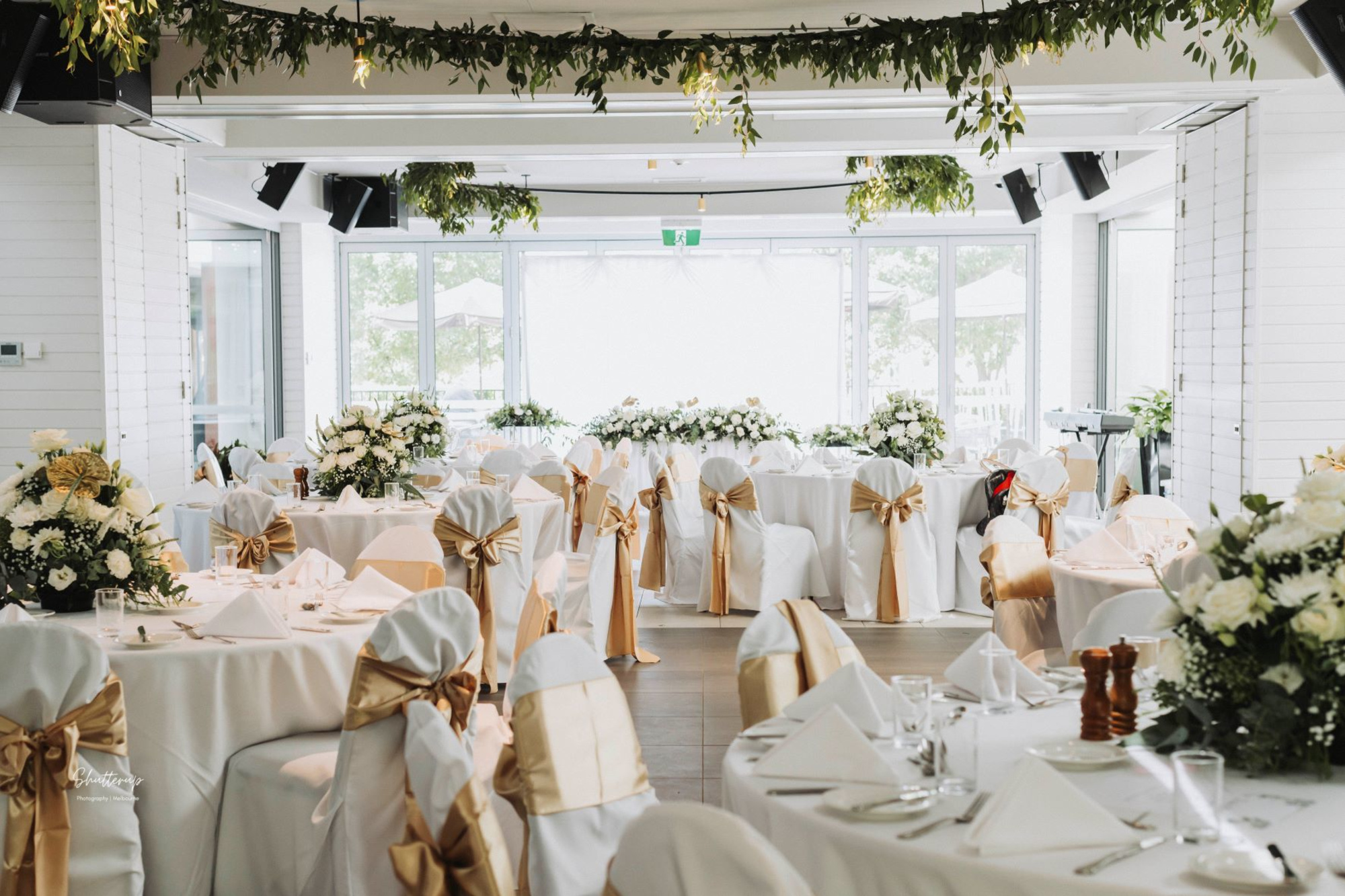 Elegant indoor wedding reception with white and gold decor, floral centerpieces, and greenery hanging from the ceiling.