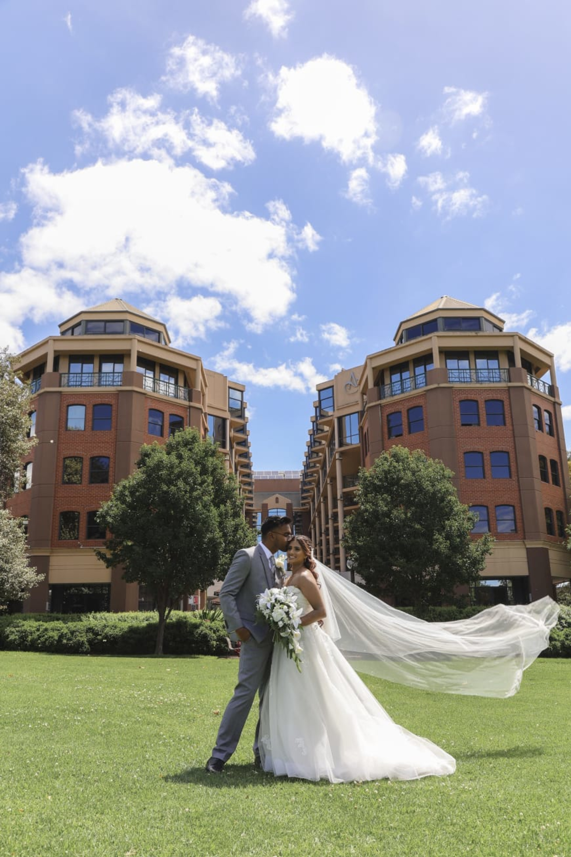 Bride and groom embrace on a green lawn with her veil flowing and tall city buildings behind them.