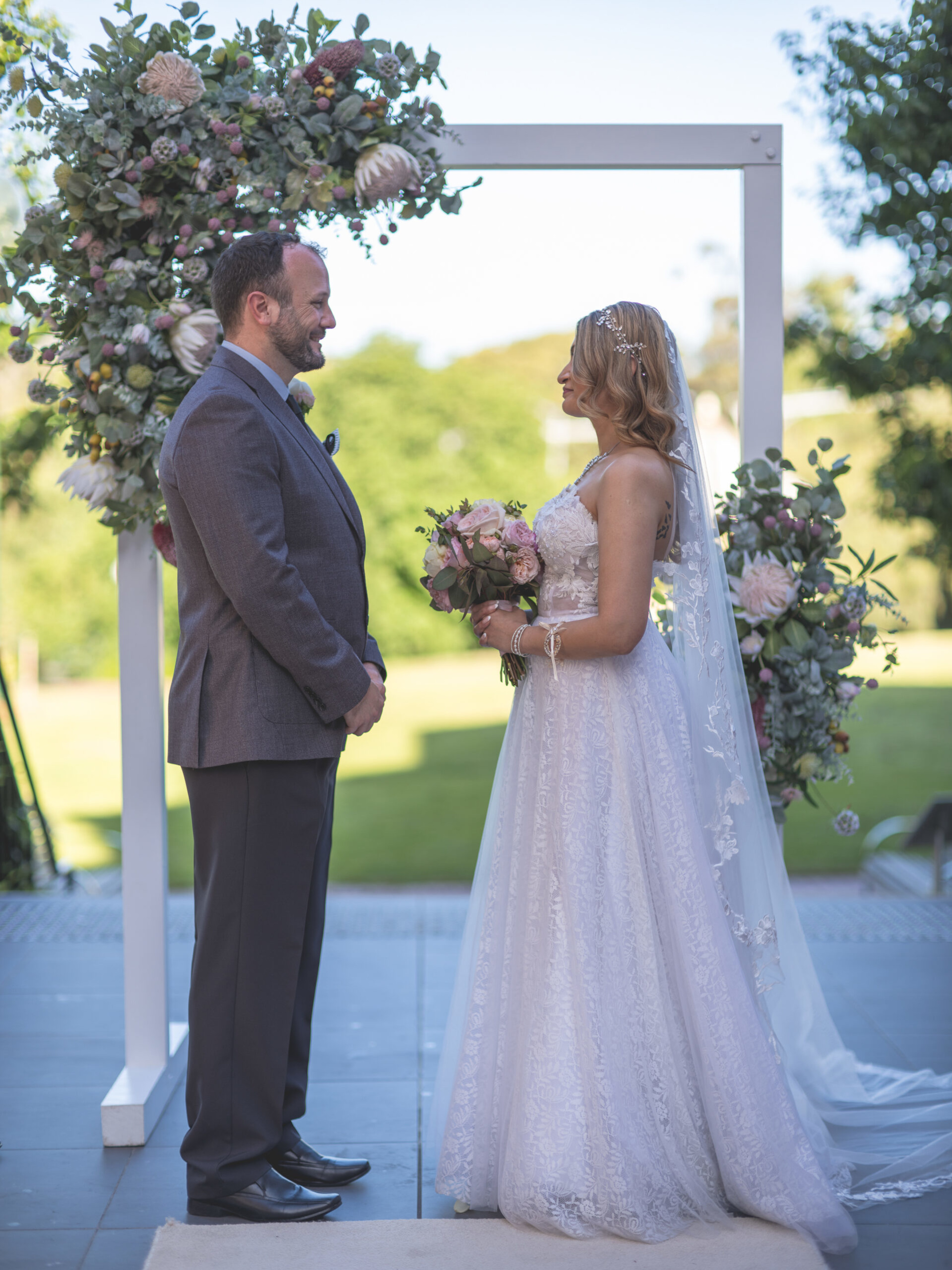 Bride and groom face each other under a floral arch during an outdoor wedding ceremony.