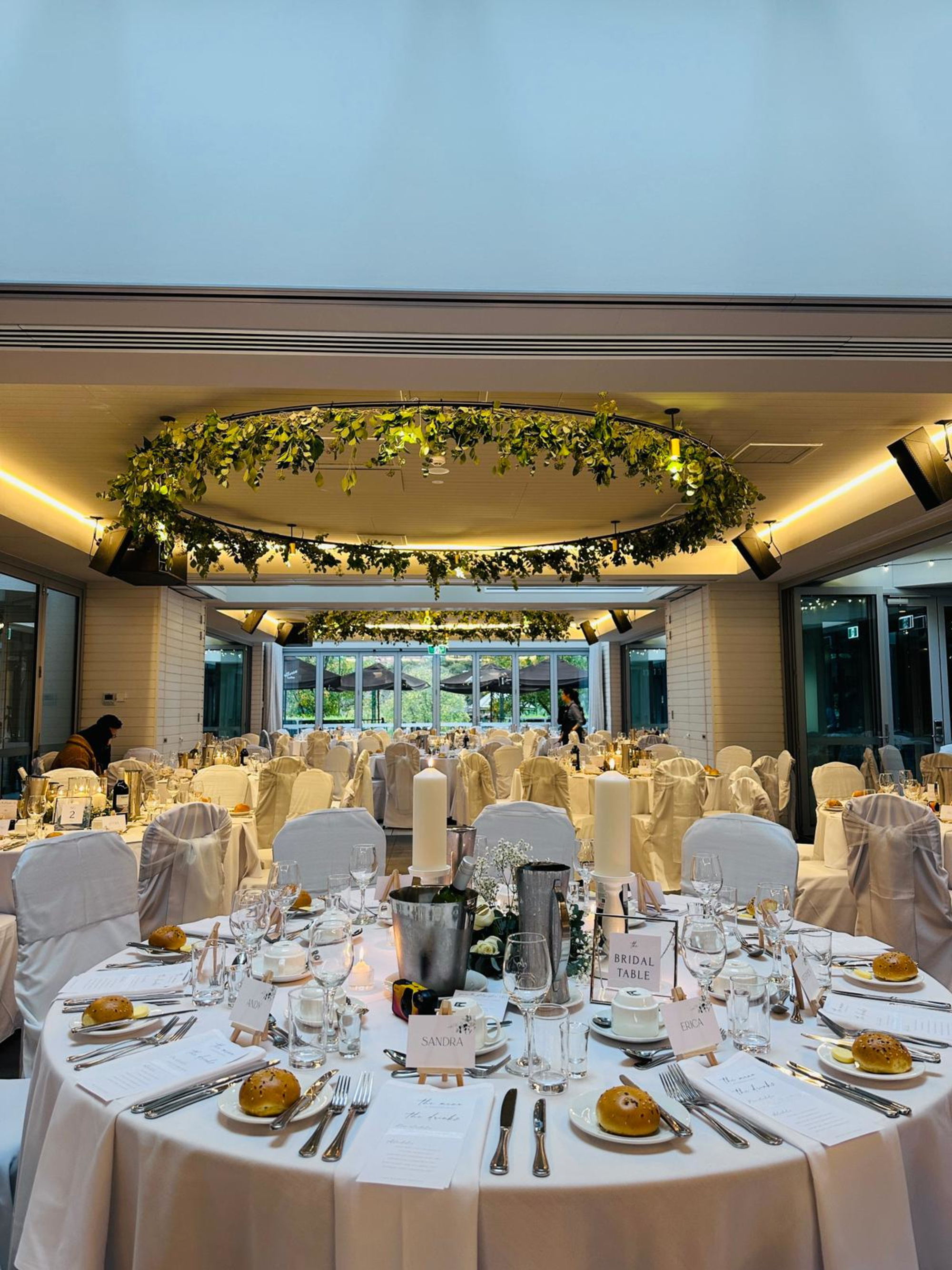 Elegant indoor wedding reception with round tables, white linens, and greenery hanging from circular ceiling fixtures.