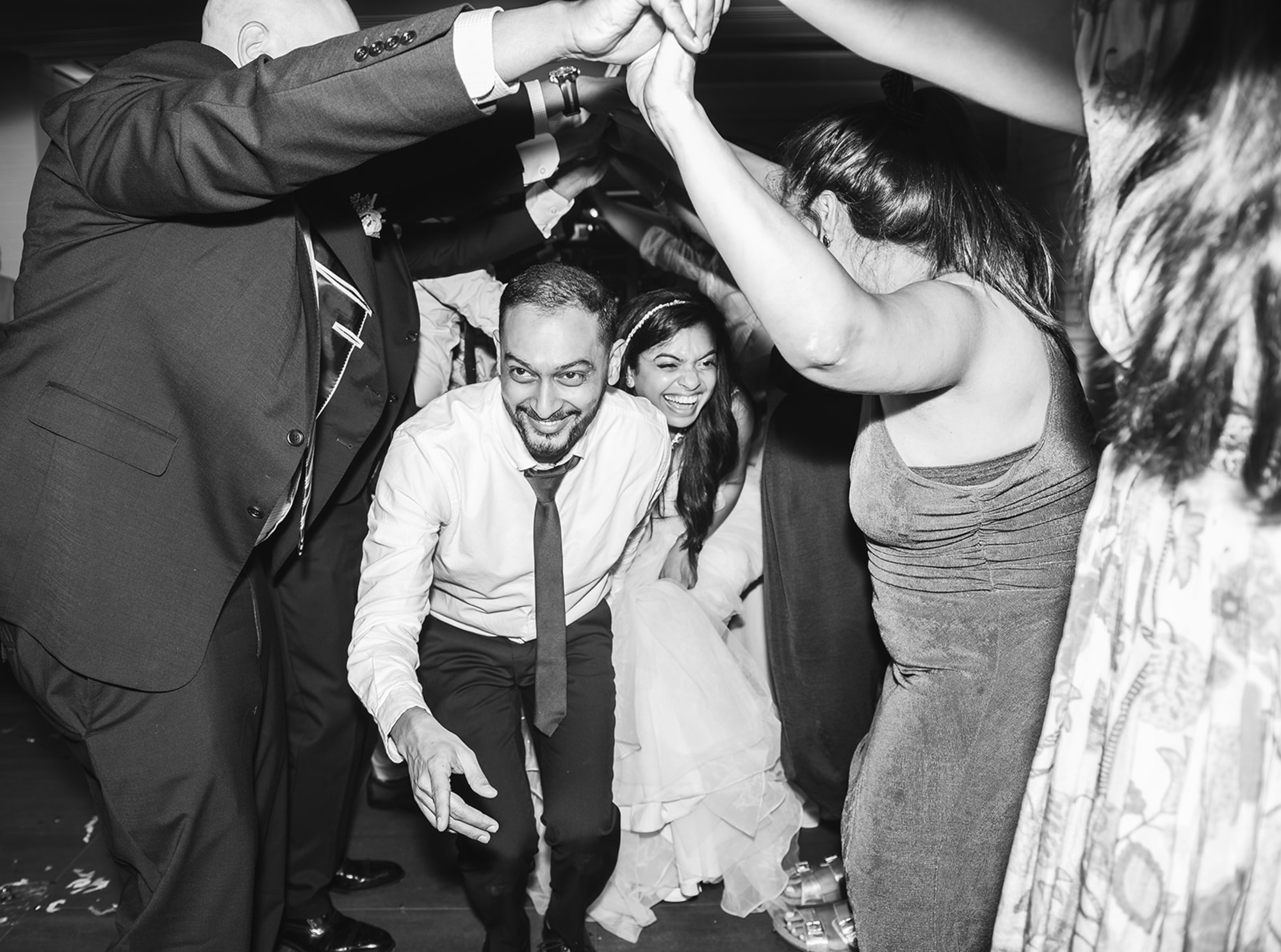 Candid black and white photo of a bride and groom laughing as they duck under guests’ raised arms during the wedding reception.