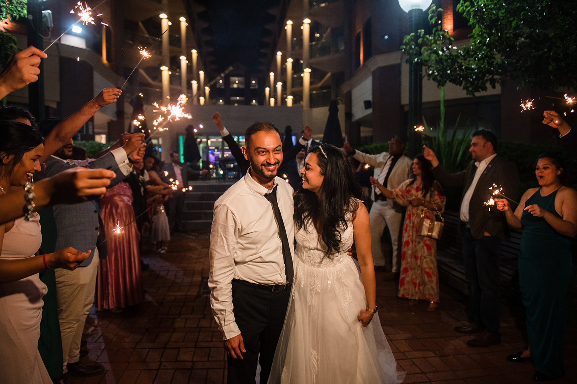 Bride and groom walk through a nighttime sparkler exit surrounded by cheering wedding guests.