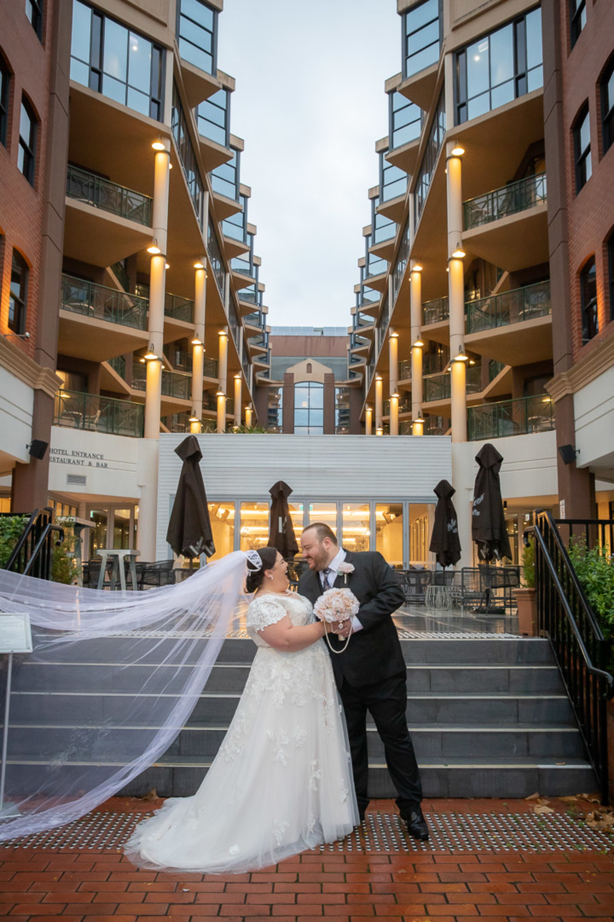 Bride and groom embrace on city steps between modern buildings with a long veil flowing behind them.