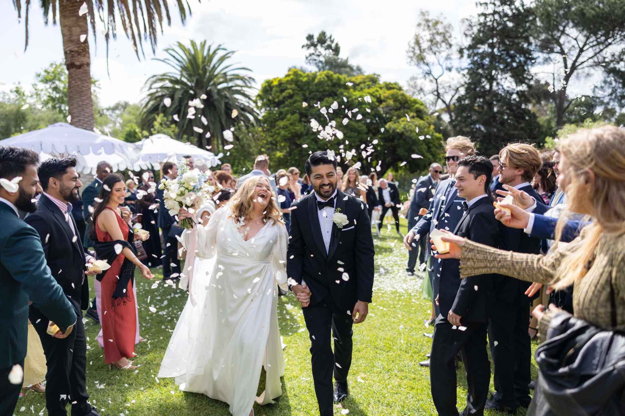 Bride and groom walk through guests tossing flower petals at an outdoor garden wedding.
