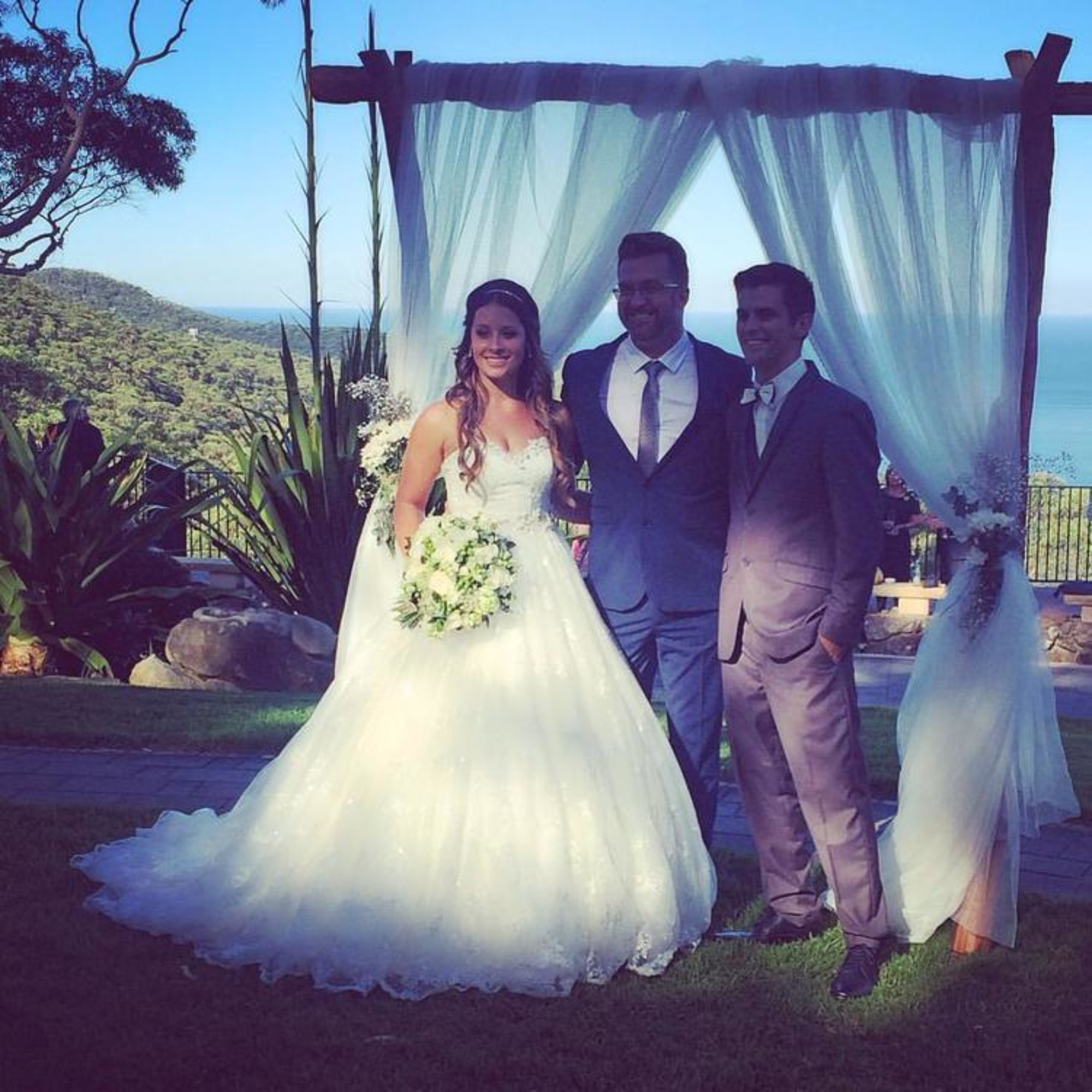 Bride and groom pose with an officiant under a draped wooden arch overlooking a scenic ocean and hillside view.