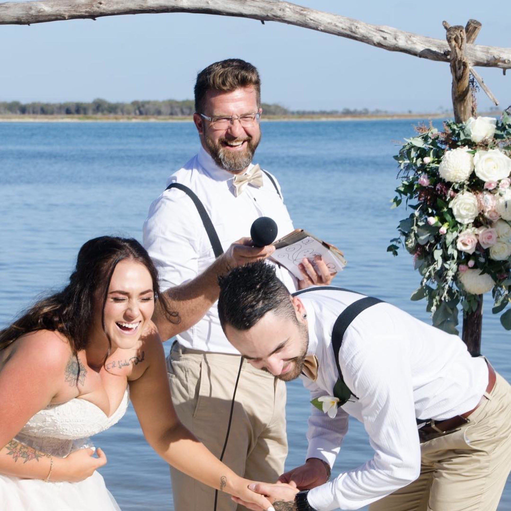Laughing bride and groom share a lighthearted moment with their celebrant during a sunny beachside wedding ceremony.