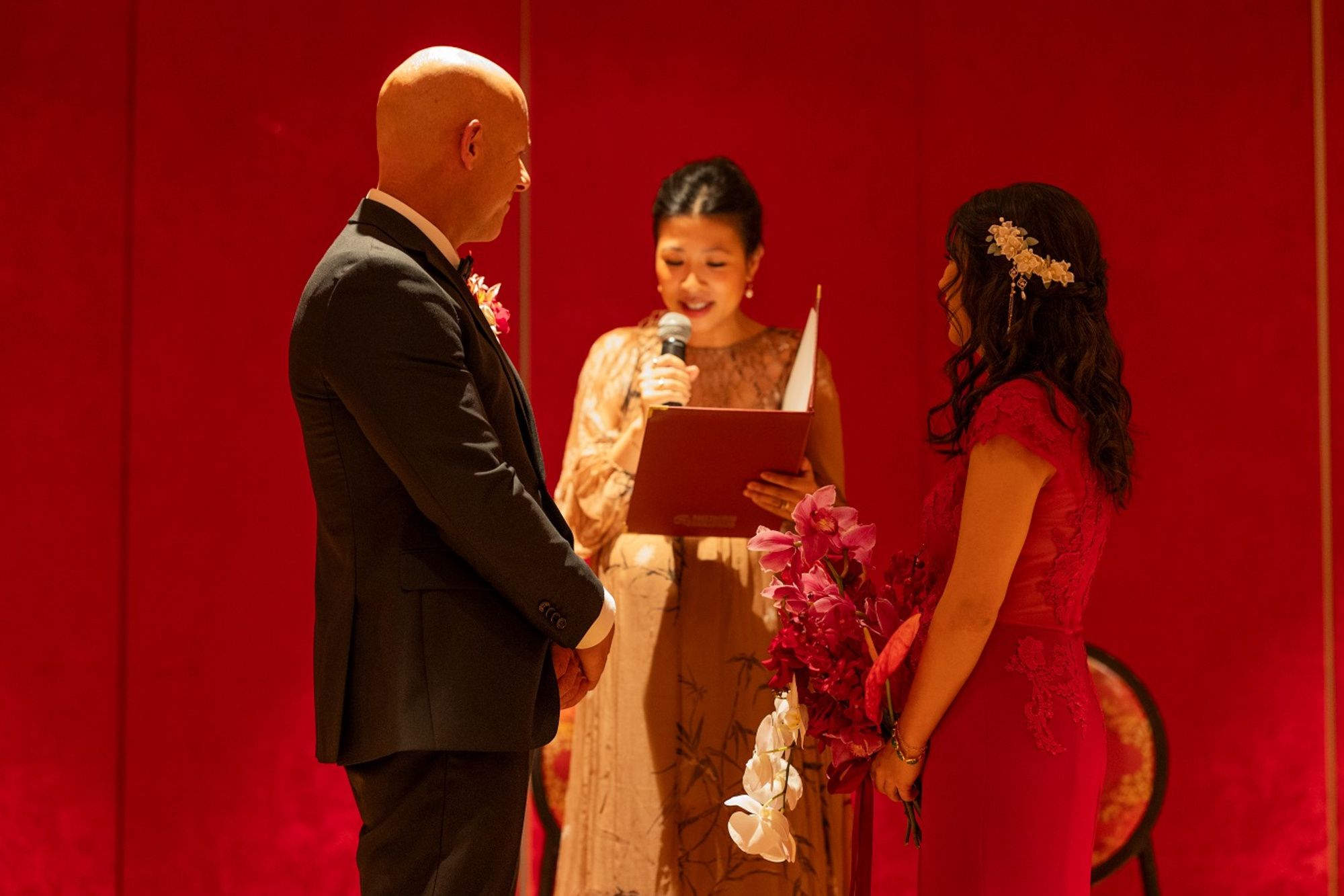 Couple stands before an officiant during an intimate indoor wedding ceremony with a bold red backdrop.