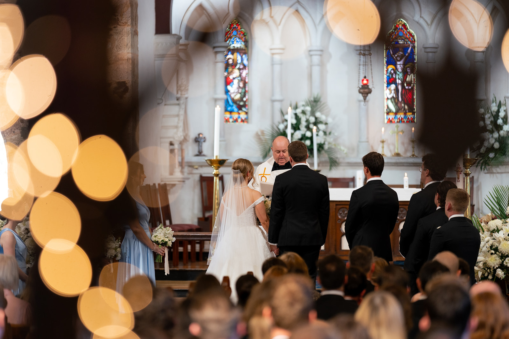 Bride and groom stand at the altar during a church wedding ceremony framed by warm bokeh lights.